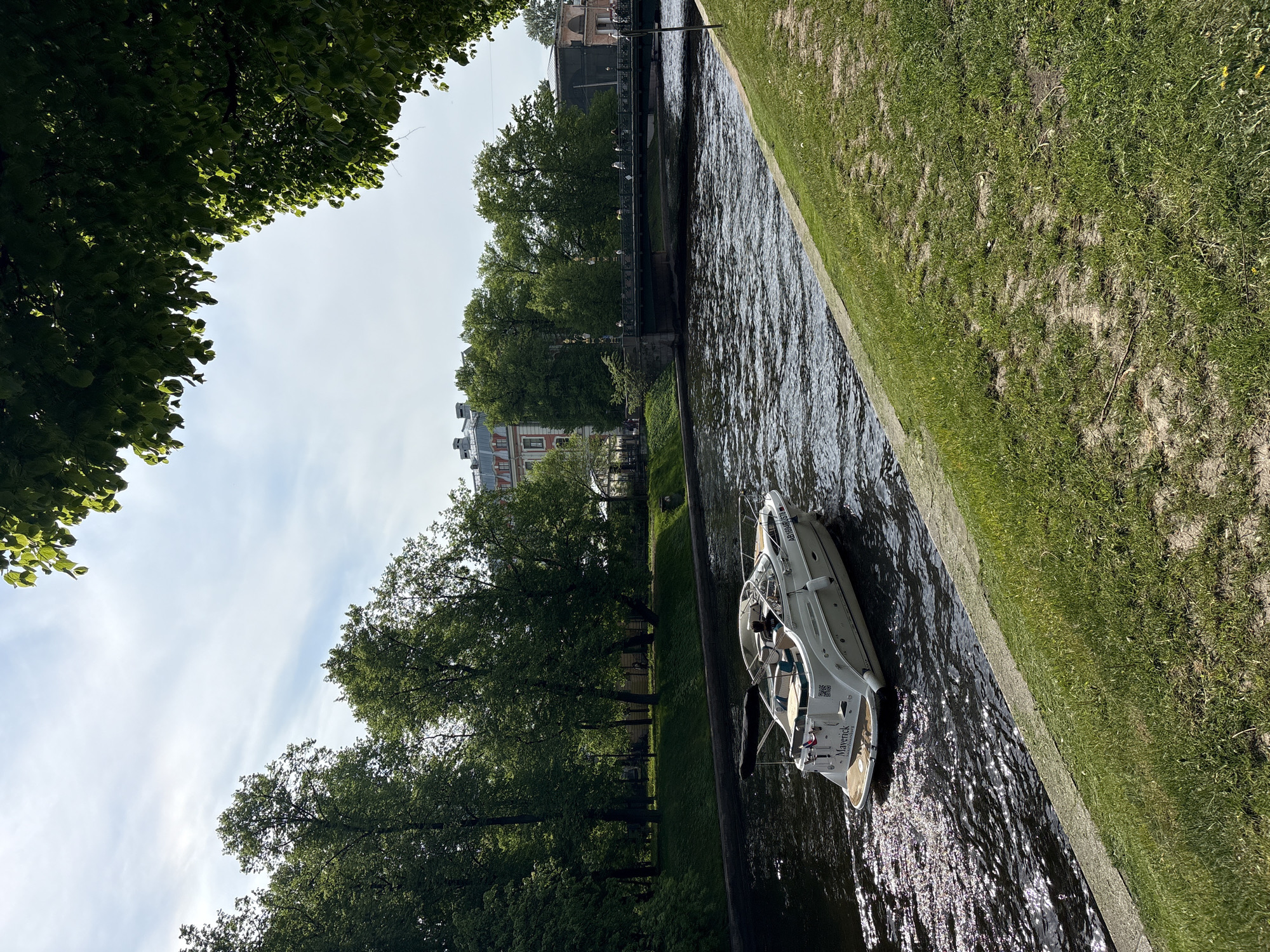 The image depicts a boat navigating a canal in Saint Petersburg, Russia. The canal is lined with stone embankments and grassy banks, with trees providing shade along the water's edge. Buildings are visible in the background, suggesting an urban setting. The boat, a small white motorboat, is occupied by at least one person. The waterway appears relatively narrow, typical of the canals that traverse the city. The lighting indicates it is daytime.