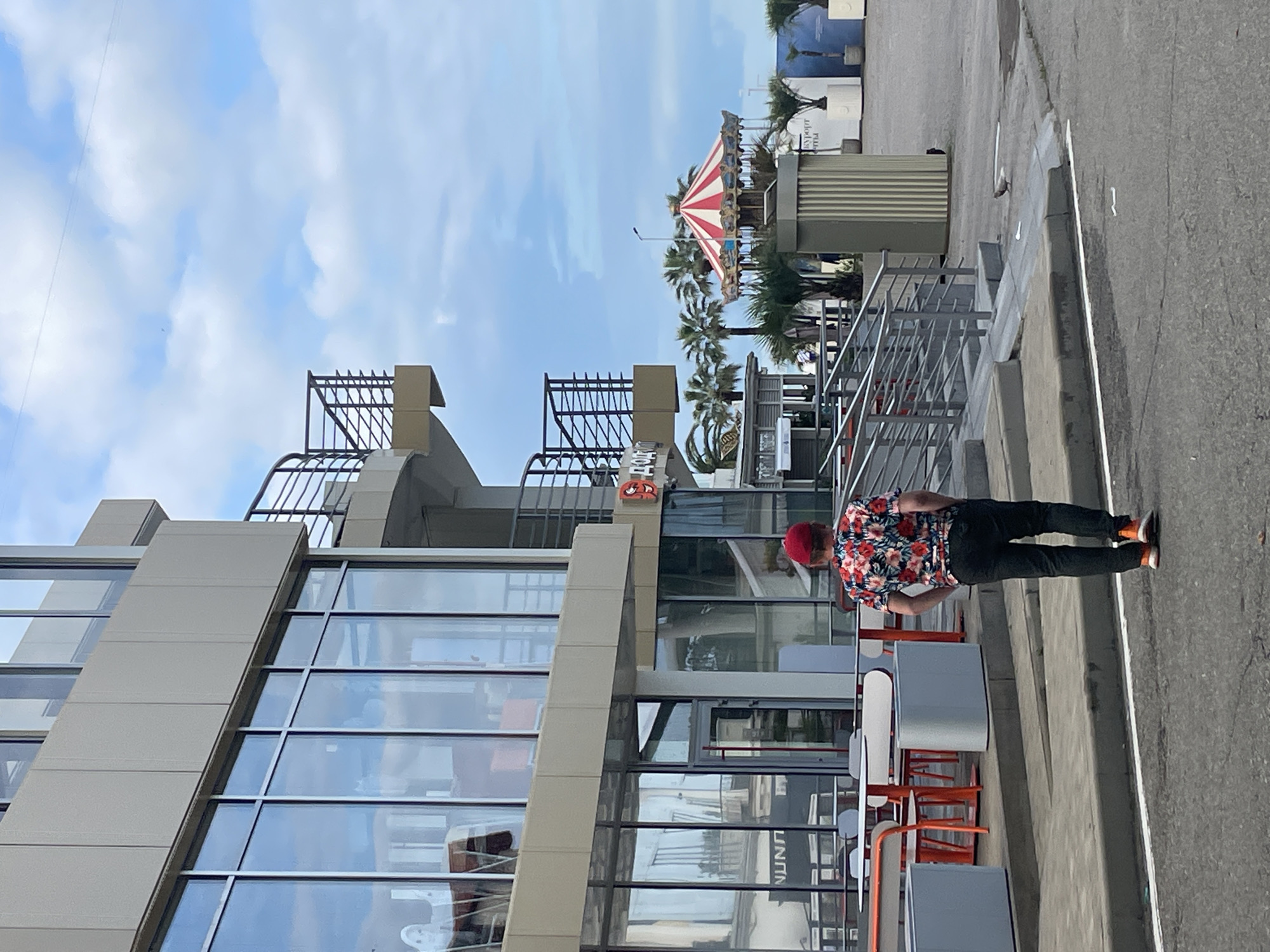 The image depicts an outdoor scene in Sochi, Russia, possibly near the coast given the palm trees and bright sky. In the foreground, a man wearing a red floral shirt, black pants, and orange sneakers walks away from the camera, facing towards a modern building with large glass windows. The building appears to be a commercial establishment, with outdoor seating and tables visible in front. Beyond the building, a carousel with red and white stripes is visible, along with more palm trees and white planters. The overall atmosphere suggests a public space, potentially a promenade or park area in Sochi.