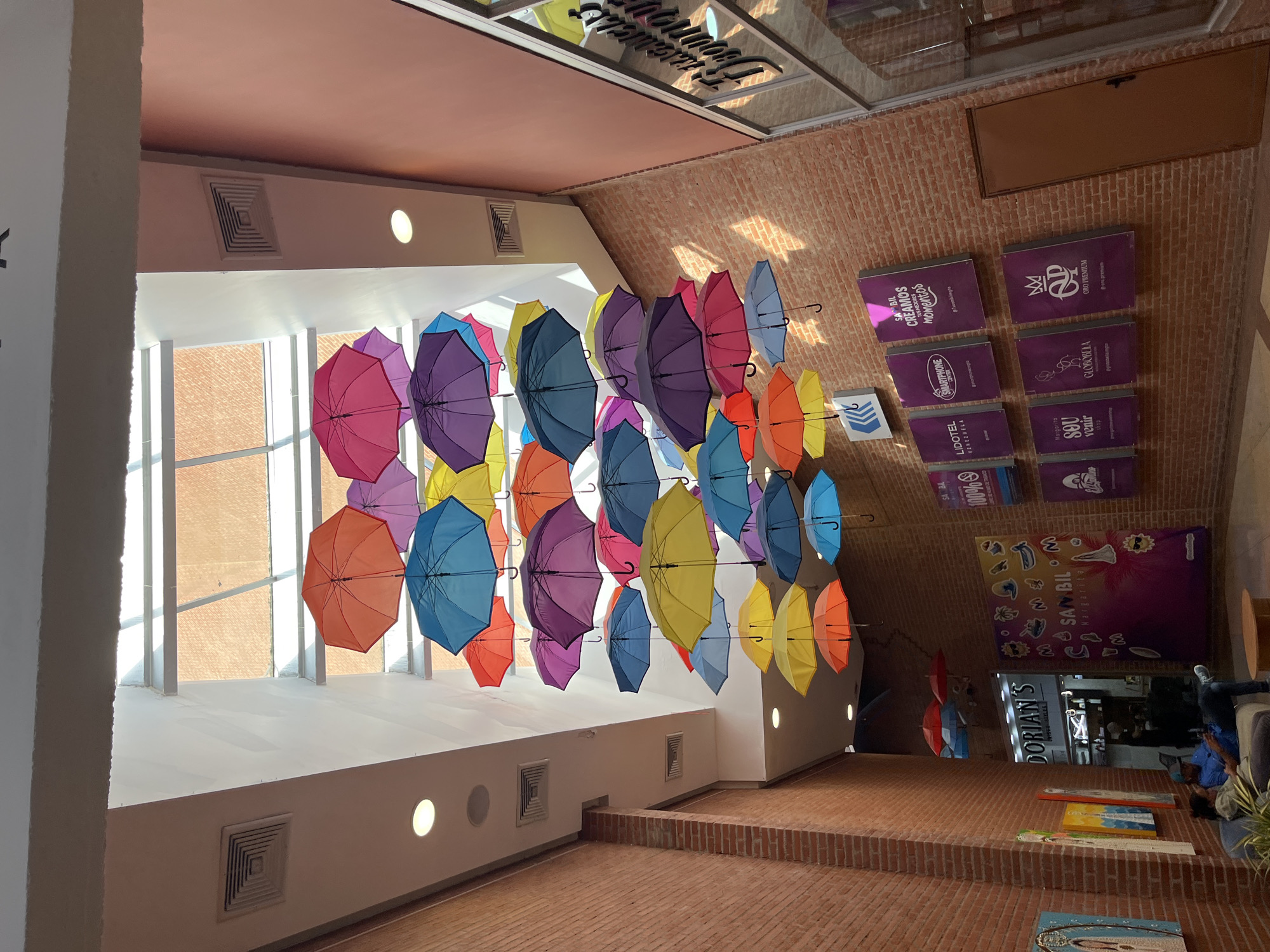 An overhead view captures a vibrant indoor commercial space, likely a shopping mall in Pampatar, Venezuela, adorned with numerous colorful umbrellas suspended from the ceiling. The ceiling, a bright white structure, features large windows on one side, allowing natural daylight to illuminate the area. Below the ceiling, a large array of open umbrellas in various hues including pink, purple, blue, yellow, orange, and red are densely hung, creating a striking decorative display.
The right side of the image features a prominent brick wall, against which several purple rectangular signs are mounted, displaying text and logos for various businesses. Among these are "ORO PREMIUM @oro_premium," "GLOBOBELA," "LIDOTEL venezuela," "SMARTPHONE CENTER," and a partially visible "SA BIL CREAMOS TUS MEJORES momentos." A larger banner on the brick wall reads "SAMBIL Margarita." Further down, a shopfront with the name "DORIAN'S" is visible.
The floor is made of reddish-brown brick tiles, with a raised section also constructed from brick. In the lower right portion of the image, a few individuals are visible, one person sitting on a dark bench or sofa, and another partially seen nearby, suggesting people are relaxing or waiting. The bright, natural lighting indicates it is daytime. The overall ambiance is lively and inviting, enhanced by the playful umbrella decor.