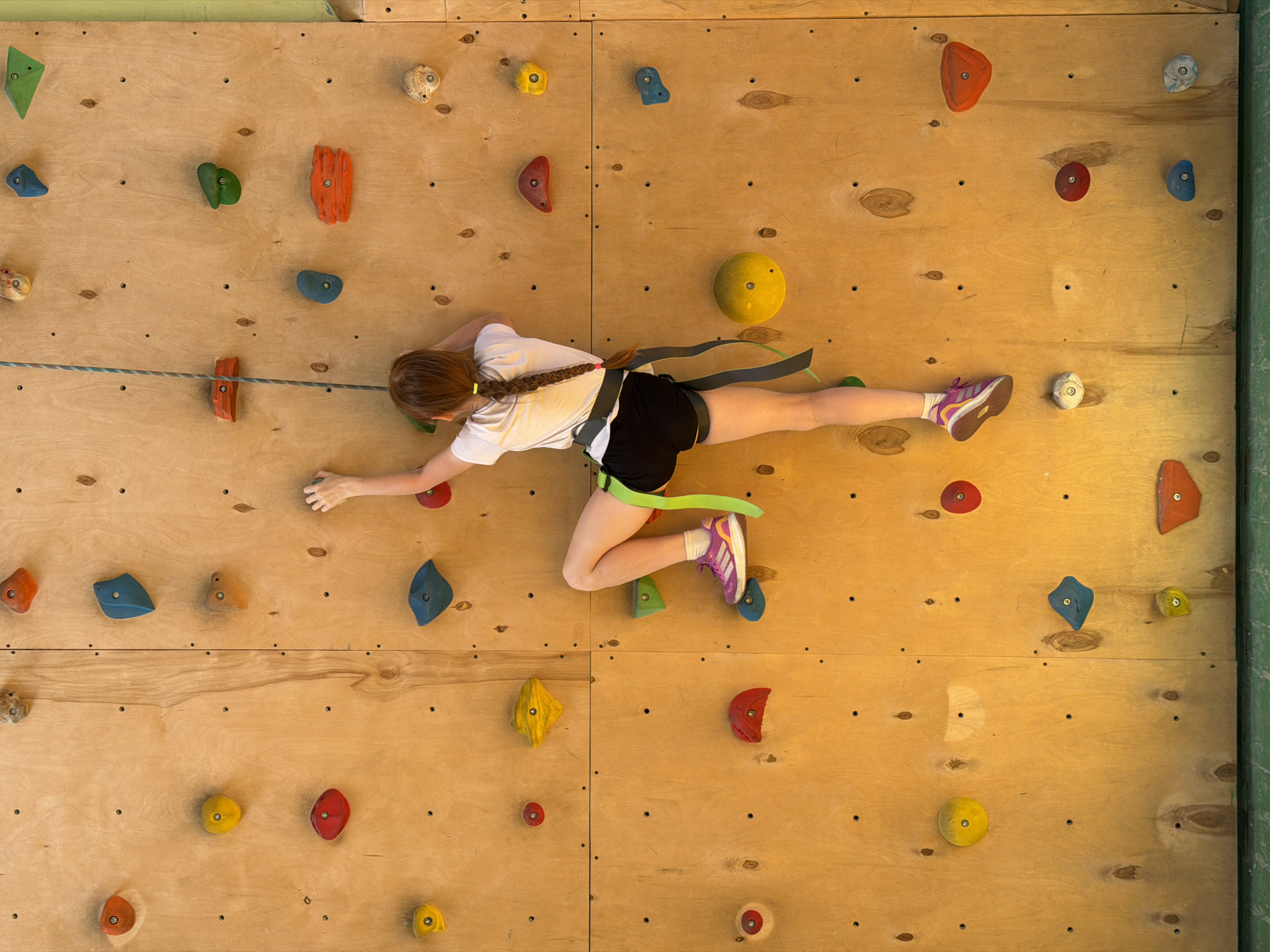 A young girl with a braided ponytail is suspended on a climbing wall. She wears a white t-shirt, black shorts, a climbing harness, and athletic shoes. The climbing wall is made of light-colored wood and has various colored climbing holds attached to it. She is attached to a rope that appears to be secured to the top of the wall. The wall is indoors. The image does not provide enough information to determine the city of Izhevsk, Russia.