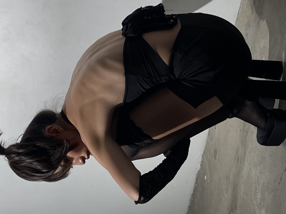 The image depicts an indoor scene in the city of London, United Kingdom. A single individual, appearing female, is captured in a crouching posture, facing left. The individual has dark hair styled in an elevated bun with bangs, and wears a black choker and an ear piercing. Attire consists of a black strapless garment with back ruching and a textured embellishment, black lace fingerless opera gloves on both forearms, black sheer thigh-high stockings, and black platform shoes with block heels. The subject's back is partially visible, showing contours of the spine and shoulder blades. The background is a plain, light-colored wall, and the floor is a grey, textured concrete surface. Lighting appears to originate from the upper right, casting shadows on the subject and floor.