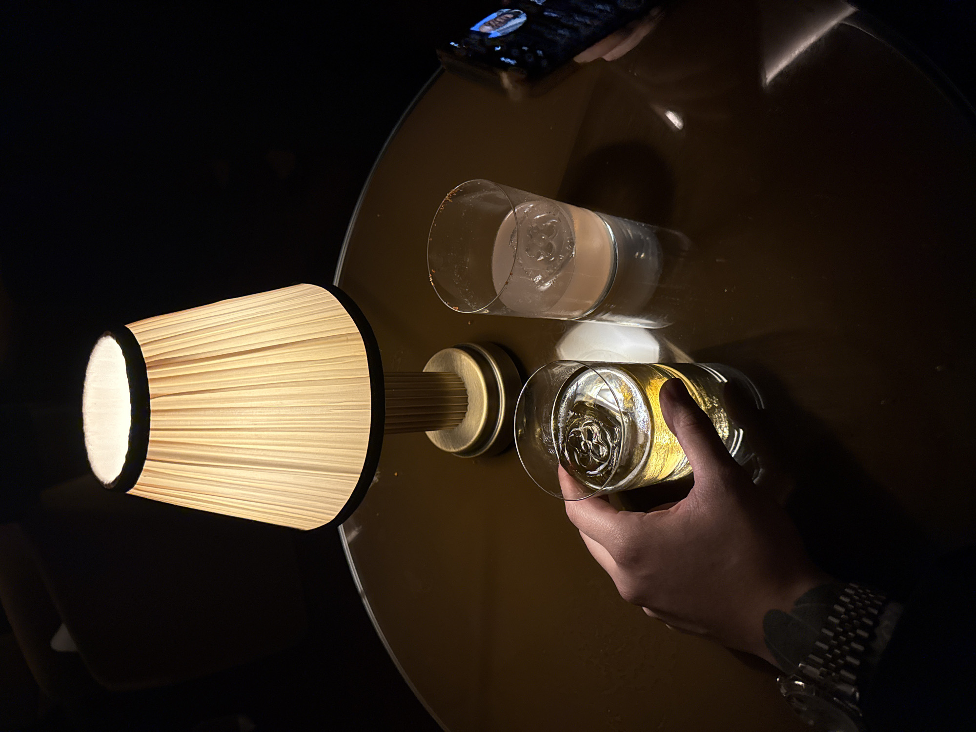 The image depicts a table scene, likely in a dimly lit establishment in London, United Kingdom. The glossy round table surface reflects light, showcasing two cylindrical glasses: one containing a light-colored beverage, held in a hand adorned with a watch and a dark tattoo, and the other appears to hold a clearer drink with ice. A lampshade with a pleated texture emits a warm glow, its light casting shadows on the table. The top corner reveals a person holding a smartphone. The overall atmosphere suggests a modern and dimly lit bar or lounge setting.
