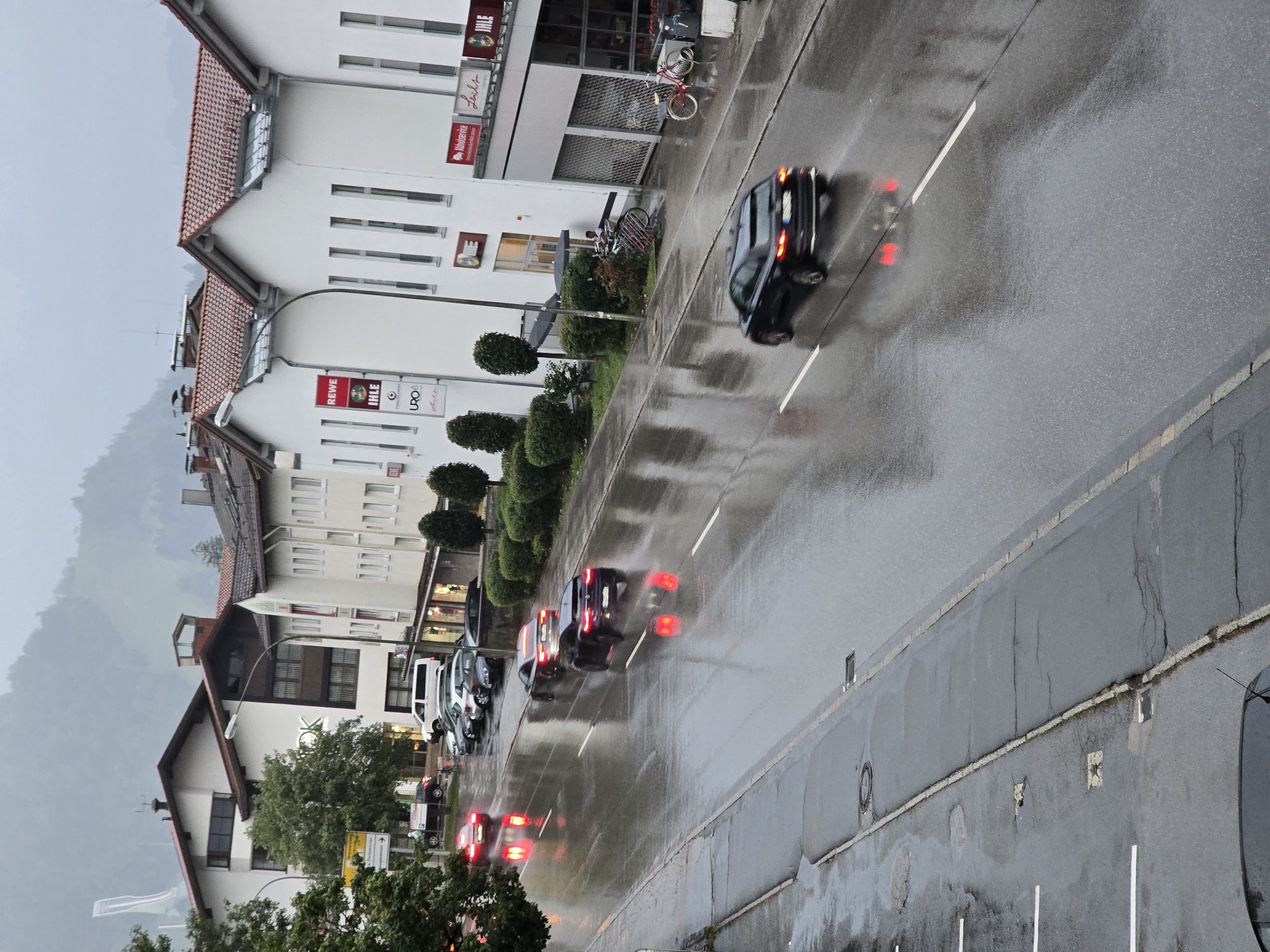 The image captures a rainy street scene in Garmisch-Partenkirchen, Germany. The perspective is from an elevated position, looking down a wet road lined with buildings. Several cars are driving on the slick, reflective asphalt, their headlights and taillights creating streaks of light. The buildings on the right side of the road appear to be commercial establishments with signage. In the background, the overcast sky and hazy mountains suggest a typical Bavarian weather day. The overall atmosphere is one of a quiet, functional town on a day with inclement weather.