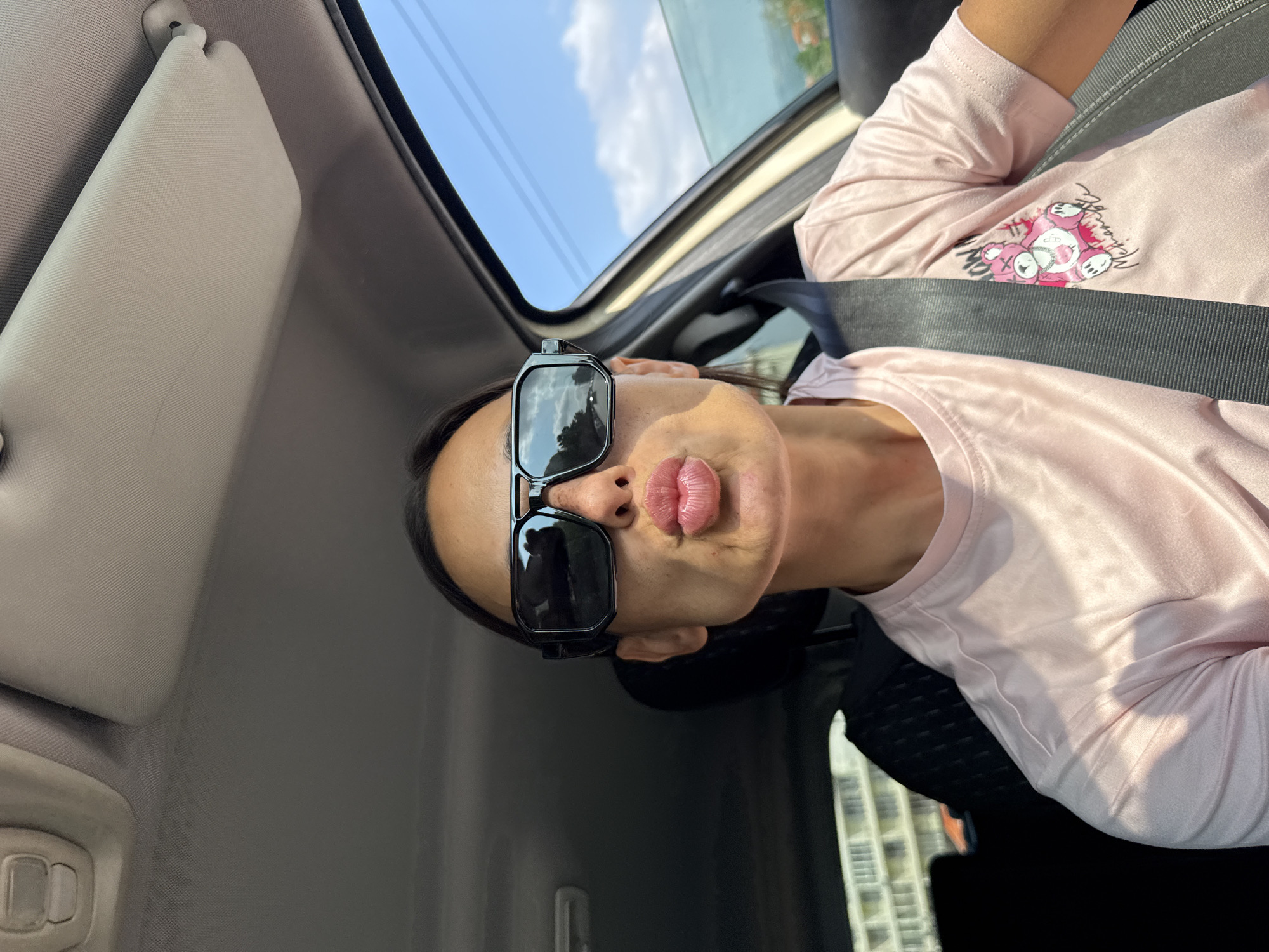 A woman is taking a selfie in a car. She is wearing sunglasses and a pink t-shirt with a graphic of a teddy bear. She is pouting her lips, as if to blow a kiss. The car appears to be in motion, as indicated by the reflection of the road and scenery in her sunglasses. The background shows a sunny sky with some clouds, and out-of-focus buildings and greenery. The woman is wearing a seatbelt. The interior of the car is visible, including the sun visor and part of the dashboard. There is no specific indication of the location in Pleven, Bulgaria.