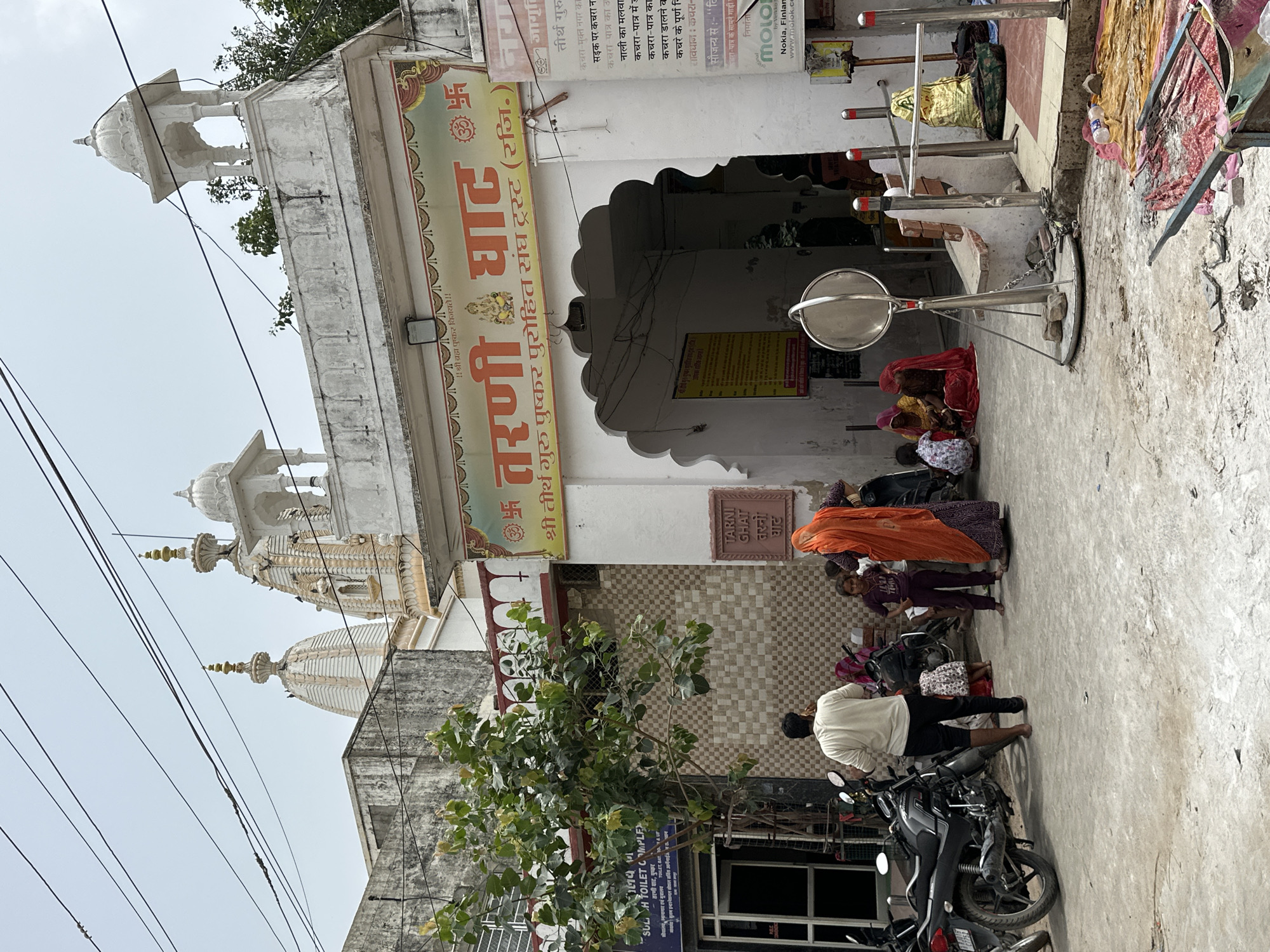 The image depicts an outdoor scene in Pushkar, India, likely in front of a religious establishment. In the foreground, a light-colored paved area shows signs of wear and tear, with scattered debris and a small cart loaded with fabric. Several motorcycles are parked on the left side of the frame.
A group of people are visible, including a man in a white shirt and dark pants standing with his back to the camera. Near him, a young girl in a purple outfit and a woman in an orange sari are present. Further back, several other women in colorful saris are seated on the ground.
The background features a large, ornate archway leading into a building. Above the arch, a sign with Hindi text and religious symbols is prominently displayed. The architecture includes multiple domes and decorative elements typical of Indian temples. To the left of the archway, a tree grows, partially obscuring a blue sign with text in both English and Hindi. The overall environment suggests a bustling public space associated with a place of worship.