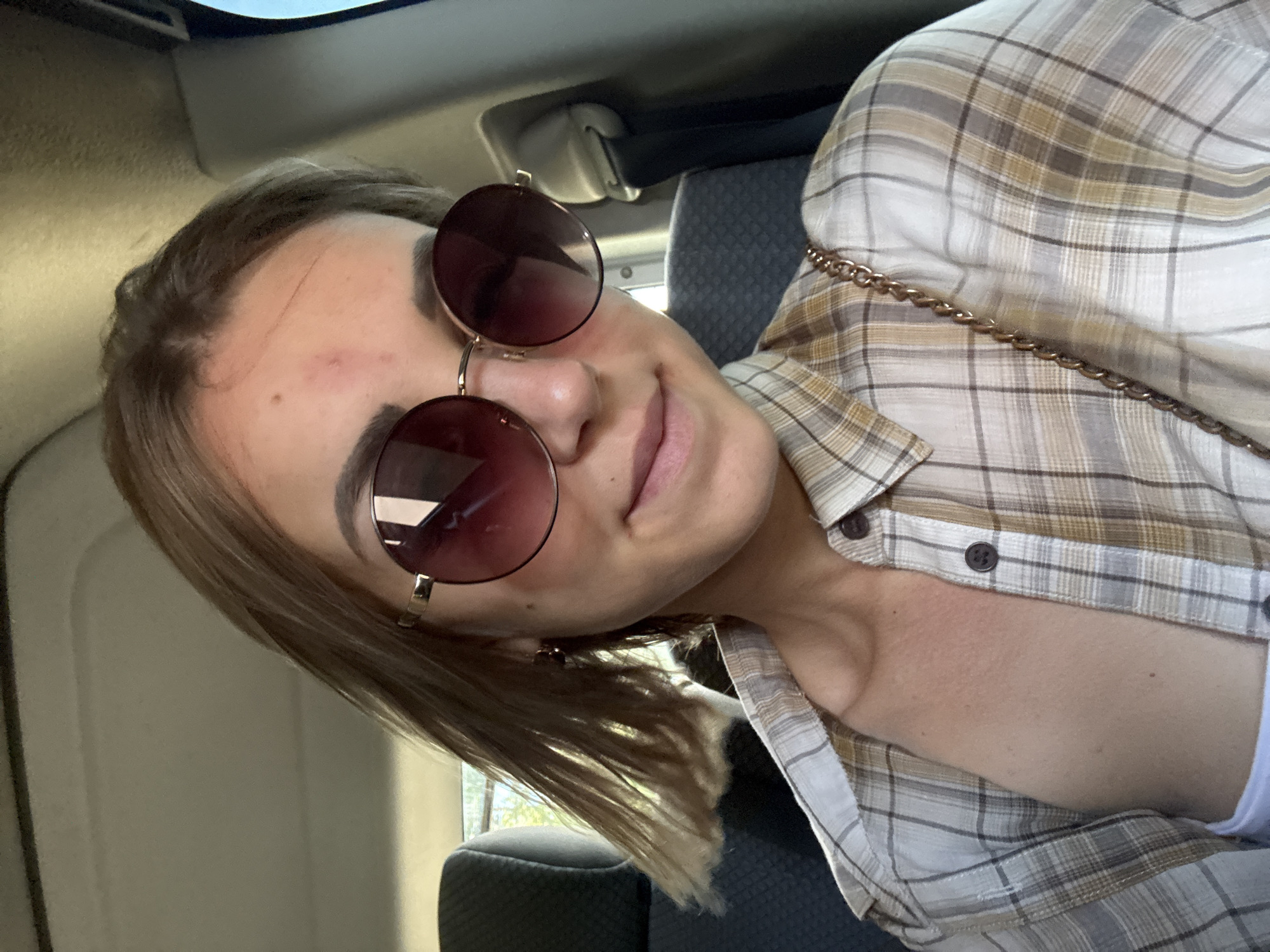 A young woman is captured from the chest up, seated in the back of a vehicle. She is wearing round, oversized sunglasses with dark lenses and a plaid shirt that is unbuttoned, revealing a white undergarment. Her hair is light brown and falls around her shoulders, appearing to be slightly windswept. The interior of the car is visible behind her, with car seats and a seatbelt. The lighting suggests it is daytime, with natural light filtering in. There are no discernible urban or natural landmarks visible that would indicate a specific location within Uspenskoye, Russia. The context of the image is limited to the interior of a vehicle.