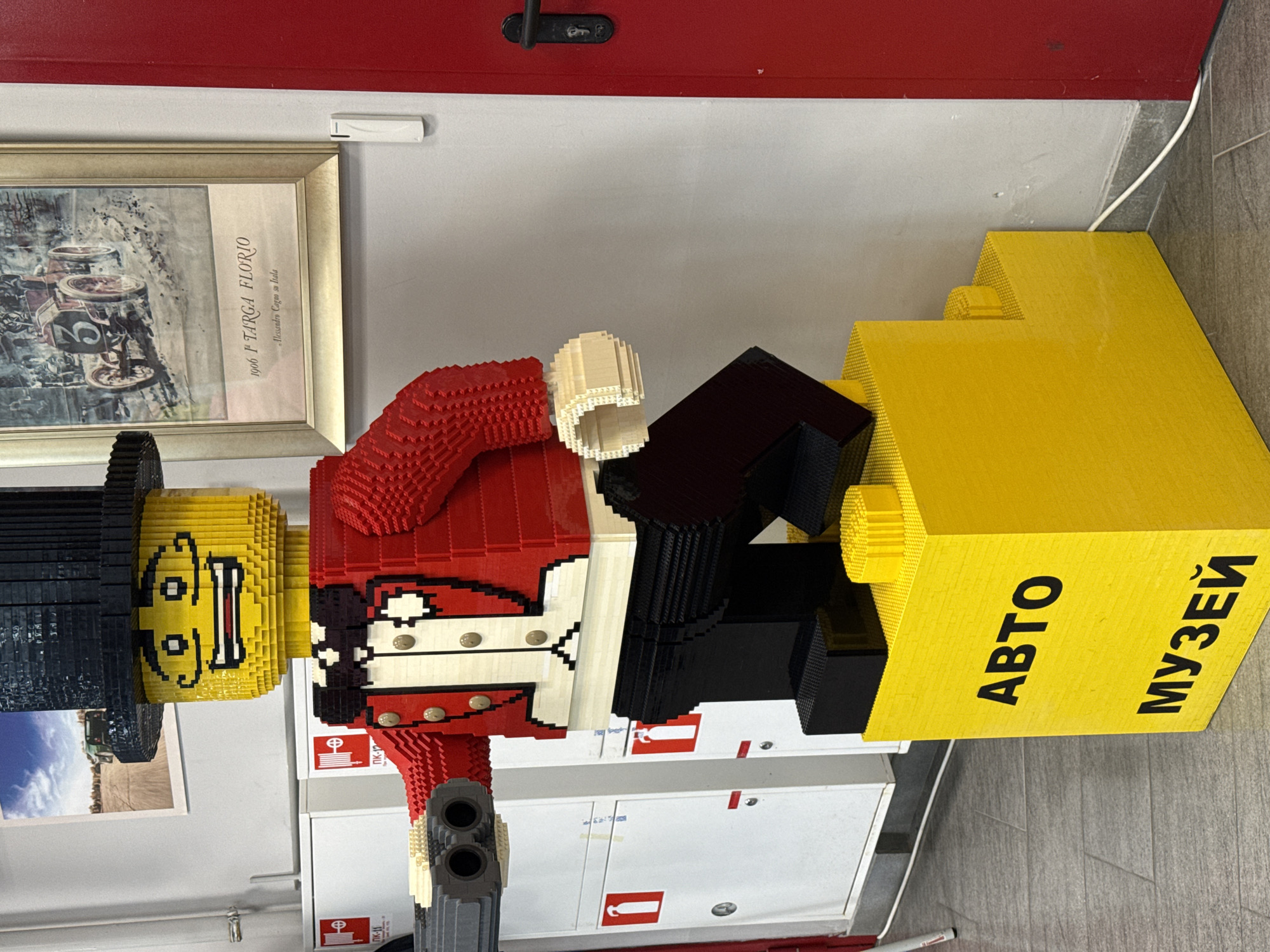 A large, colorful Lego-style figure dominates the scene, depicting what appears to be a British Guardsman or a toy soldier. The figure is constructed from numerous rectangular blocks, featuring a bright yellow head with a smiling, cartoonish face and a black top hat. Its body is predominantly red, detailed with white and black elements resembling a uniform, including a white cross belt with gold buttons. The figure holds a black and gray rifle-like object. It stands next to a large, yellow block structure that serves as a display element or base.
On this yellow structure, text is visible in Cyrillic, reading "АВТО" (AVTO - Auto) and "МУЗЕЙ" (MUZEY - Museum), indicating the setting is an automobile museum, consistent with the provided context of Sirius, Russia.
In the background, a framed picture hangs on a light gray wall. The picture depicts an old racing car, with visible text on the frame stating "1906 1° TARGA FLORIO" and "Alessandro Cagna su Italia". Above the picture, a small white electronic device is attached to the wall. The upper wall is painted red, and a black door handle is visible. Below the main figure, white cabinets with small red and white fire extinguisher signs are visible. The floor is covered with gray tiles, and a white cable runs along the base of the wall near the yellow structure. The lighting suggests an indoor setting, likely daylight or artificial overhead lighting. The image captures a static display within a public space, possibly an entrance or exhibit area of an auto museum.