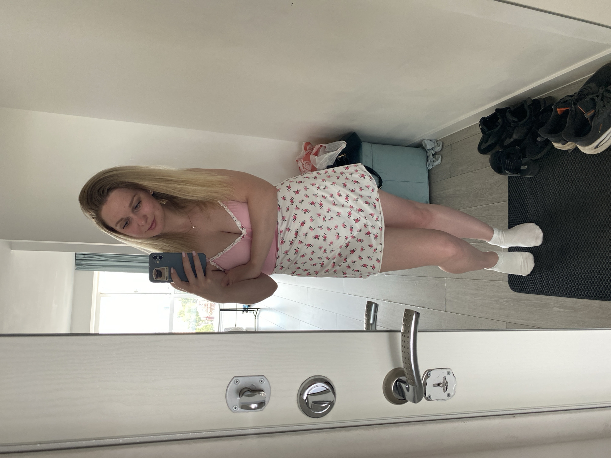 A young woman with blonde hair is taking a mirror selfie in what appears to be a hallway. She is wearing a pink camisole with white lace trim and a short white skirt with a floral print of pink flowers and green leaves. She is also wearing white ankle socks and is barefoot. Her left arm is crossed in front of her chest, and she is holding a blue smartphone in her right hand. The mirror reflects her and a portion of the room behind her, which includes a doorway with a silver handle and lock, a pale blue storage bin, some plastic bags, and a pair of black sneakers on the floor. The flooring is a light-colored wood laminate. The overall impression is that of a domestic setting, possibly an apartment or house. There is no specific information within the image that would allow for a definitive identification of the location within Izhevsk, Russia.