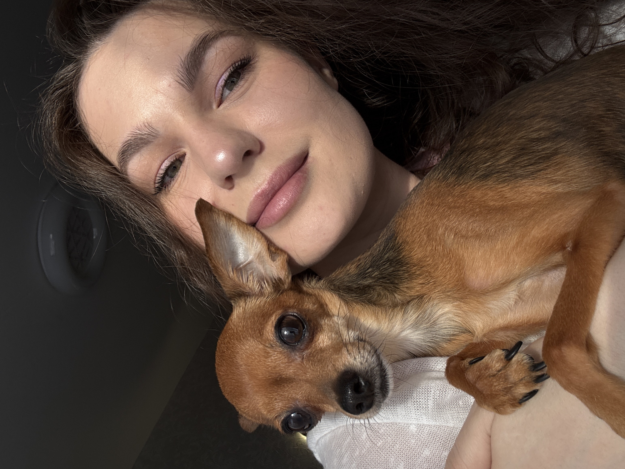 A woman is holding a small brown chihuahua-mix dog. The woman has light skin, brown hair, and is wearing pink eye shadow and lipstick. The dog is nestled against her chest. The dog has black claws. The background is a wall. The image was taken inside. There is no indication of the location being in Chaikovsky, Russia.