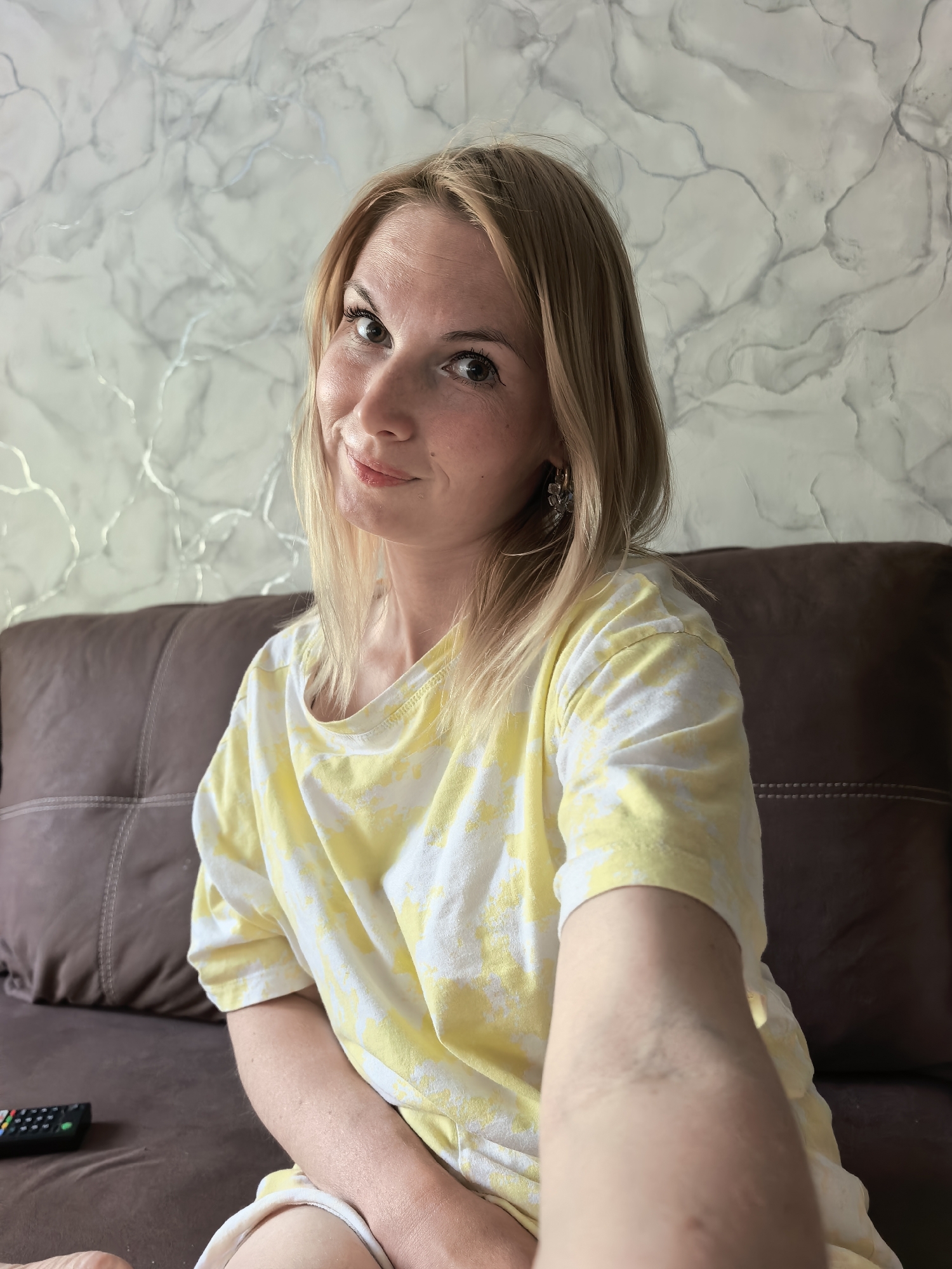 A blonde woman is sitting on a dark brown leather couch. The wall behind her is decorated with a gray and white marble pattern. The woman is wearing a yellow and white tie-dye t-shirt and has earrings. A black remote control is on the couch beside her. The image appears to be a selfie, as the woman's arm is extended towards the camera. There is no location information to confirm that the photo was taken in Sterlitamak, Russia.