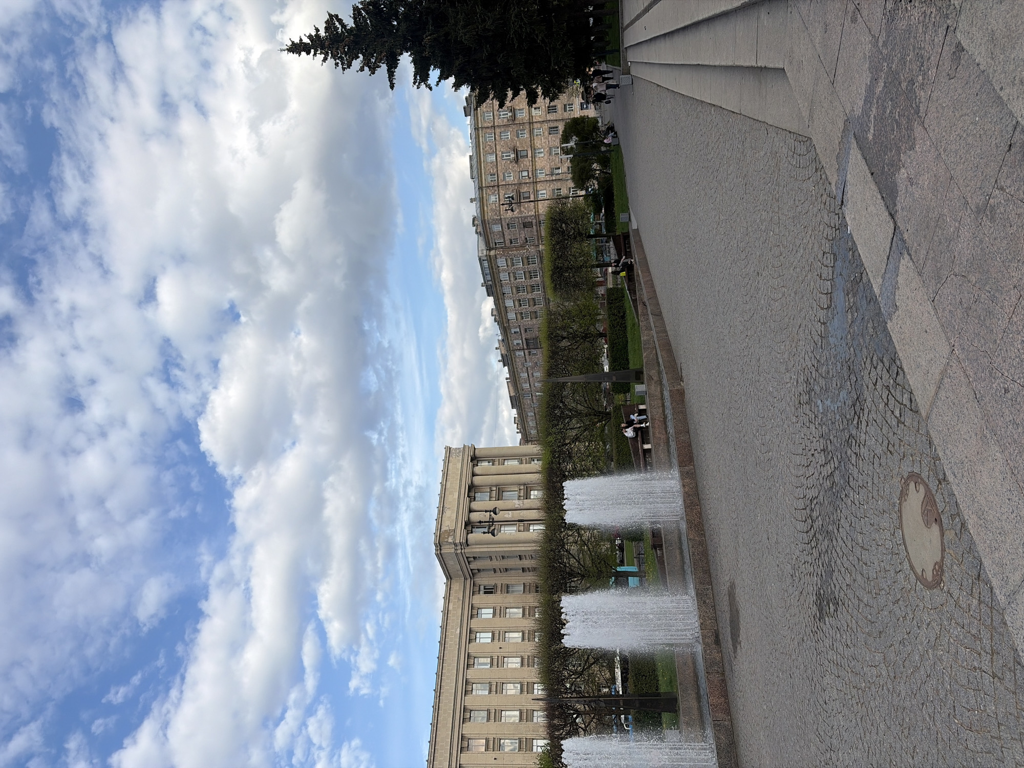 The image shows a view in Saint Petersburg, Russia, featuring a row of buildings and a fountain. The perspective is slightly rotated counter-clockwise. On the right side of the frame, there is a paved pedestrian area. In the center, a fountain with jets of water. Behind the fountain are several buildings with rows of windows. The sky is visible in the background with clouds and blue patches. The presence of people can be inferred from their locations on benches.