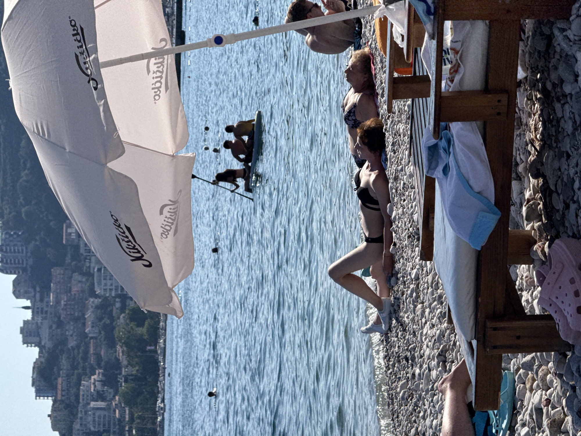 The image shows a sunny day at a pebble beach in Budva, Montenegro. The foreground is dominated by a large white umbrella, partially obscuring a view of several people. In the water, a group of individuals are engaged in recreational activities. One person is on a paddleboard, while others appear to be swimming or wading.
On the beach, a woman in a bikini is seen lounging on a sunbed. Nearby, another woman in a bikini is sitting, and an older woman in a patterned bikini is also visible. The scene is bustling with activity, suggesting a popular tourist destination. The water is a clear blue, and the sunlight reflects off its surface. The background features a hillside covered with buildings, characteristic of coastal towns in the Mediterranean.
The umbrella has text printed on it, which reads "Kušičko" and "ASBIJŠKO". The overall mood is relaxed and summery, with a sense of people enjoying a vacation. The time of day appears to be afternoon, given the angle of the sun and the shadows cast.