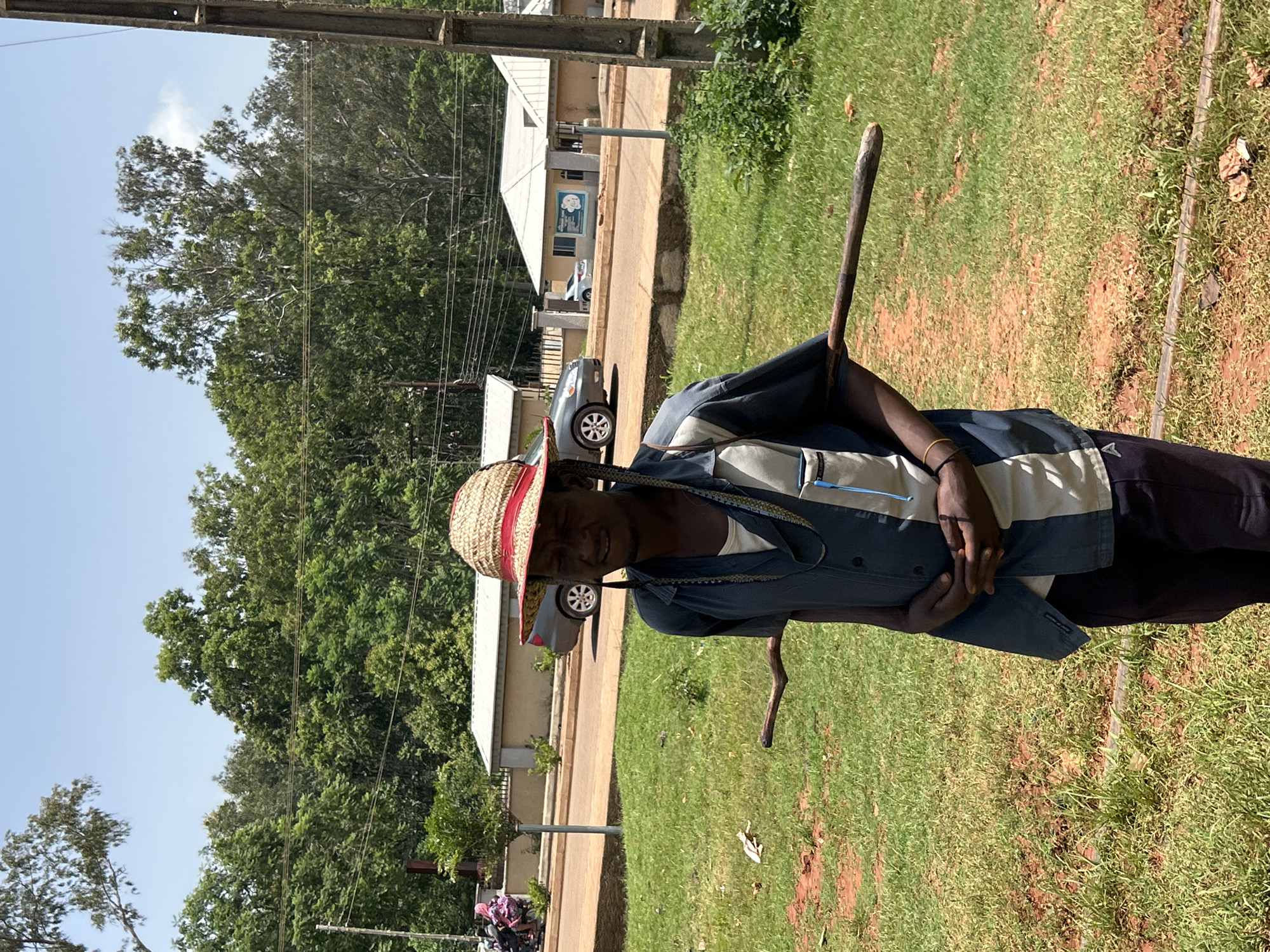 A person wearing a straw hat with a red band and a blue and white striped shirt stands outdoors in what appears to be a grassy area with some dirt patches. They are holding a long wooden stick. Behind them, a paved road is visible, with a light-colored building and some cars parked along it. Trees line the background, and overhead power lines are present. The sky is clear and blue, suggesting it is daytime and likely fair weather. The person's expression is neutral. The location is identified as Bukuru, Nigeria.