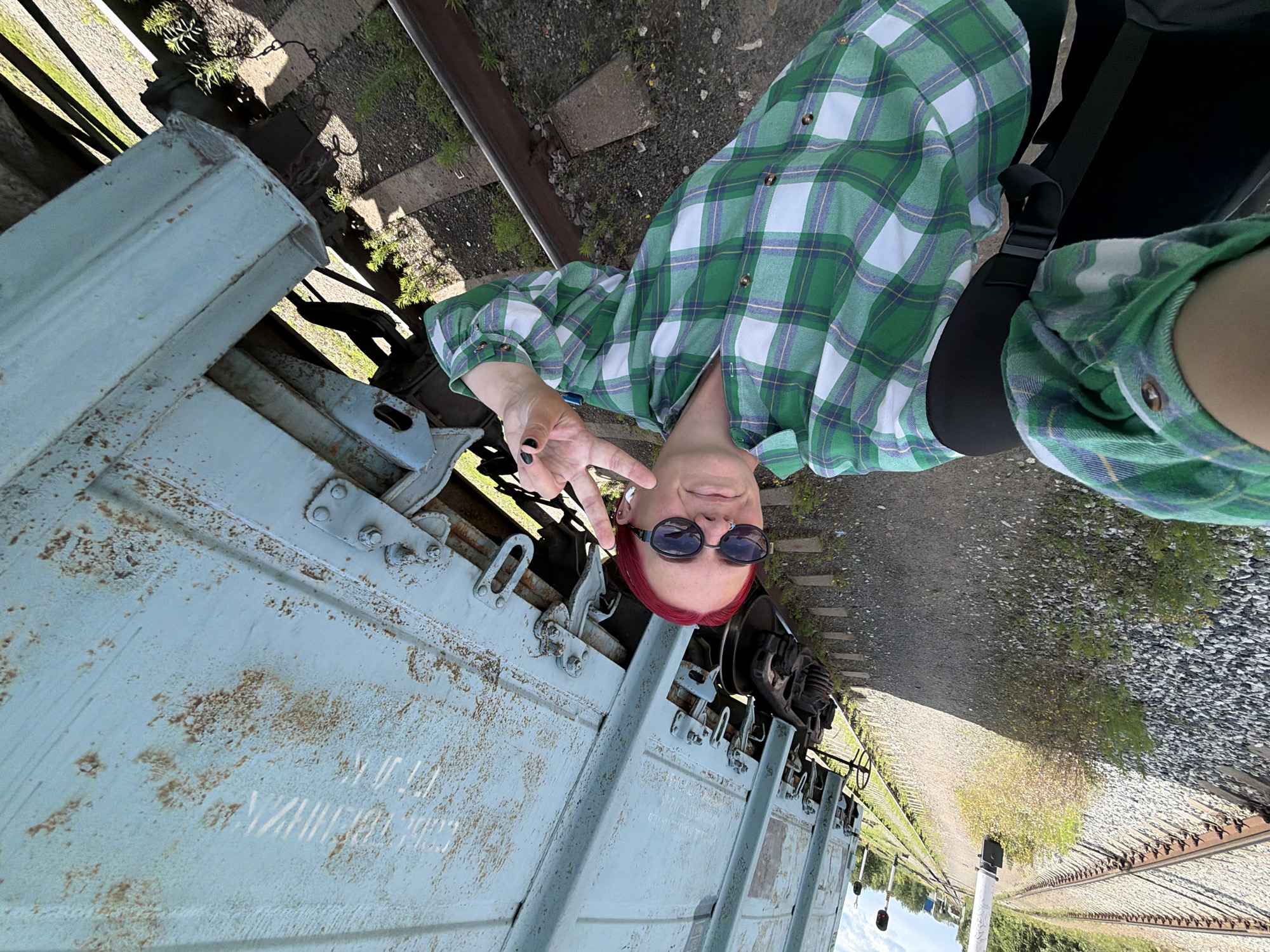 The image depicts a person with red hair, wearing sunglasses and a green and white plaid shirt, standing near a light blue train car on train tracks. The person is making a peace sign with their fingers and smiling. The scene appears to be outdoors during the day, with sunlight casting shadows. The train car is weathered, showing signs of rust and age. The background shows more train tracks, ballast, and some vegetation, suggesting an industrial or transportation-related setting. The overall impression is casual and relaxed. There is no visible text in the image.