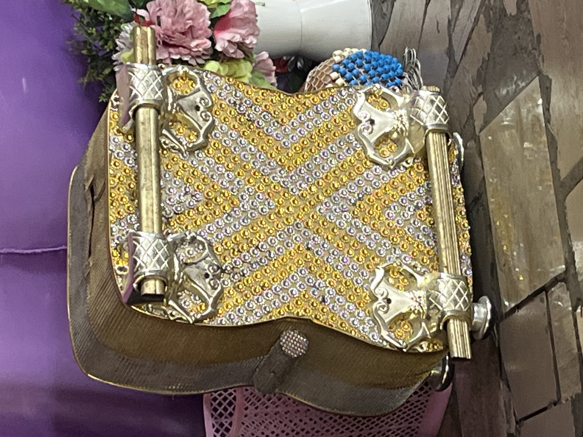 The image shows a decorated clutch purse with gold and silver gemstones, metal handle attachments, and a metallic gold body. It is positioned on top of a light purple mesh surface, likely part of a chair or ottoman. Behind the purse are flowers and part of a white object. The background includes a purple upholstered item and a wall with textured, dark gray or black tiles. There is no information to suggest that the picture was taken in Iranla, Nigeria.