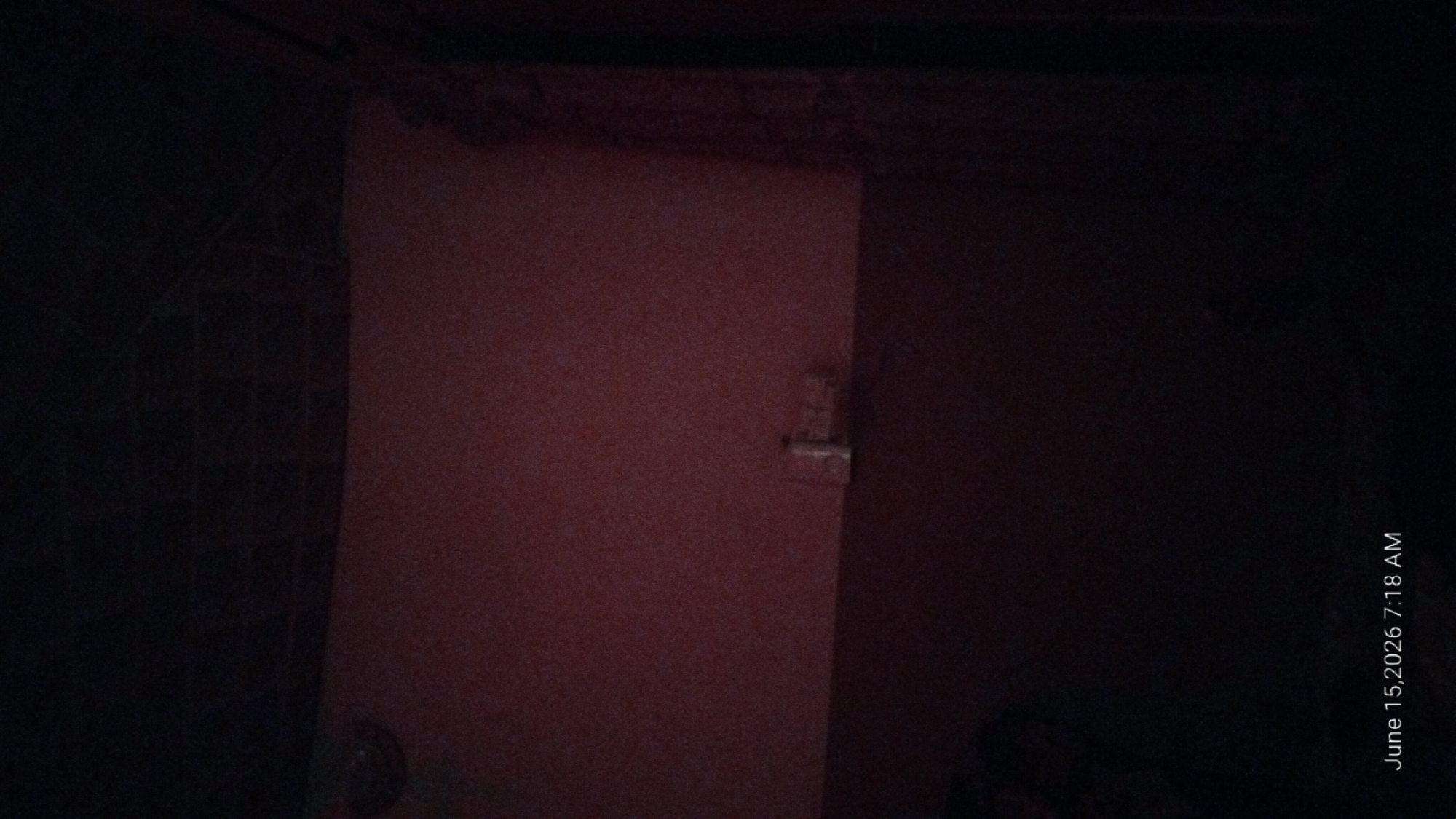 The image depicts a dimly lit room with a textured ceiling. The walls are painted in two distinct tones, with the upper portion a muted red and the lower portion a dark, almost black hue. A light switch or electrical outlet is visible on the upper wall, to the right of the center. To the far right, a dark curtain or fabric hangs. The bottom left of the image is obscured by darkness, but a sliver of a red and black patterned object can be discerned. The bottom right corner of the image displays a timestamp: "June 15, 2026 7:18 AM". The scene appears to be an interior of a residential or possibly commercial space. The location in Howayi, Nigeria, is not visually evident from the image itself.