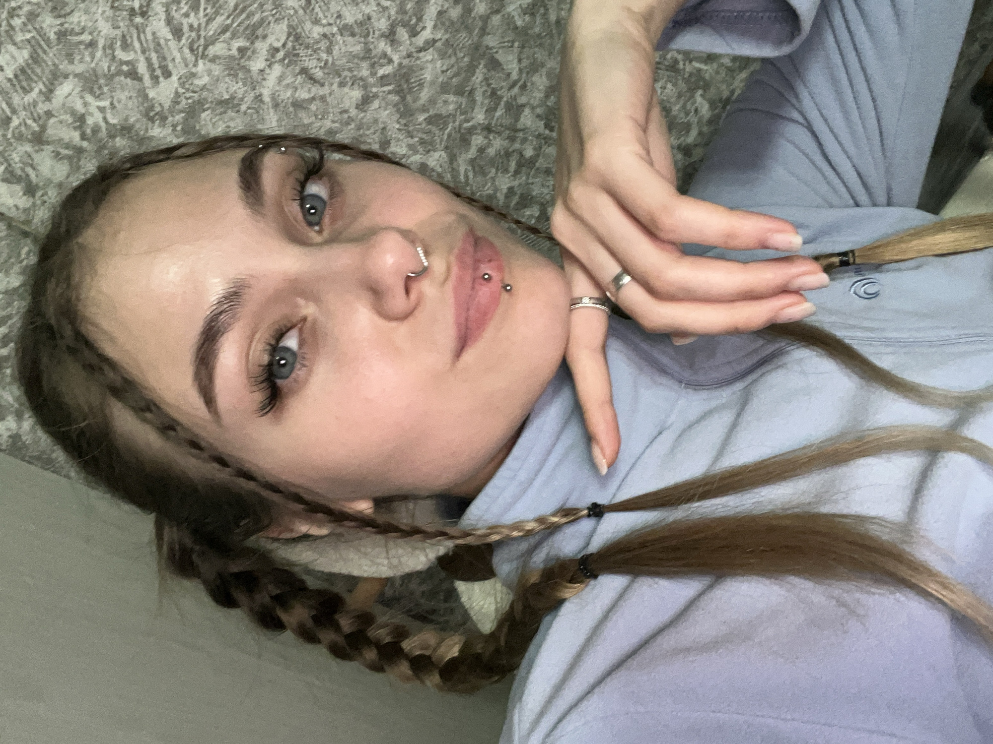 A woman is photographed from a slightly high angle, lying down with her head resting on a textured grey surface resembling a wall or bedding. She has fair skin, blue eyes, and her light brown hair is styled in braids with dark elastic bands. She has a nose piercing and a lip piercing on the right side of her mouth. Her right hand is positioned near her face, with rings on her fingers. She is wearing a light blue or lavender hooded sweatshirt with a small embroidered design on the upper right. The background behind her head is a textured grey pattern. There is no verifiable information regarding a location in Фоки, Russia.