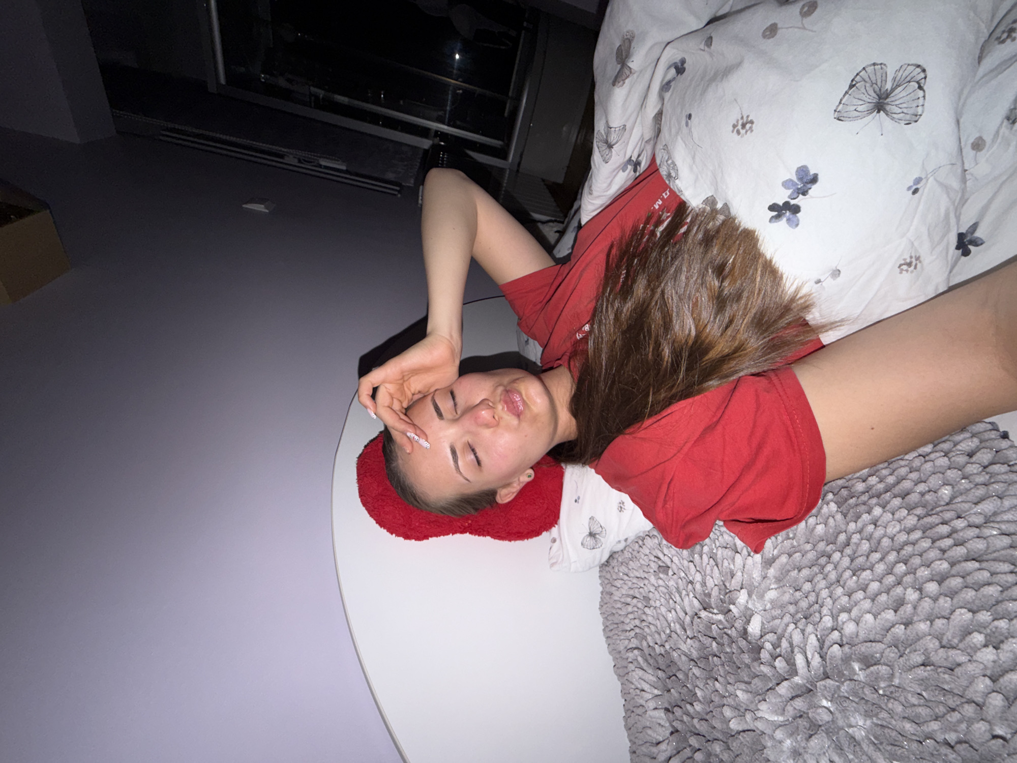 A young woman is lying on a bed. She is wearing a red shirt and has long brown hair. Her head rests on a small red pillow. The bed is covered in patterned bedding and a fluffy gray blanket. A round white table is positioned next to the bed. The room has a light purple wall. A window is visible in the background. The location is Buhovo, Bulgaria.