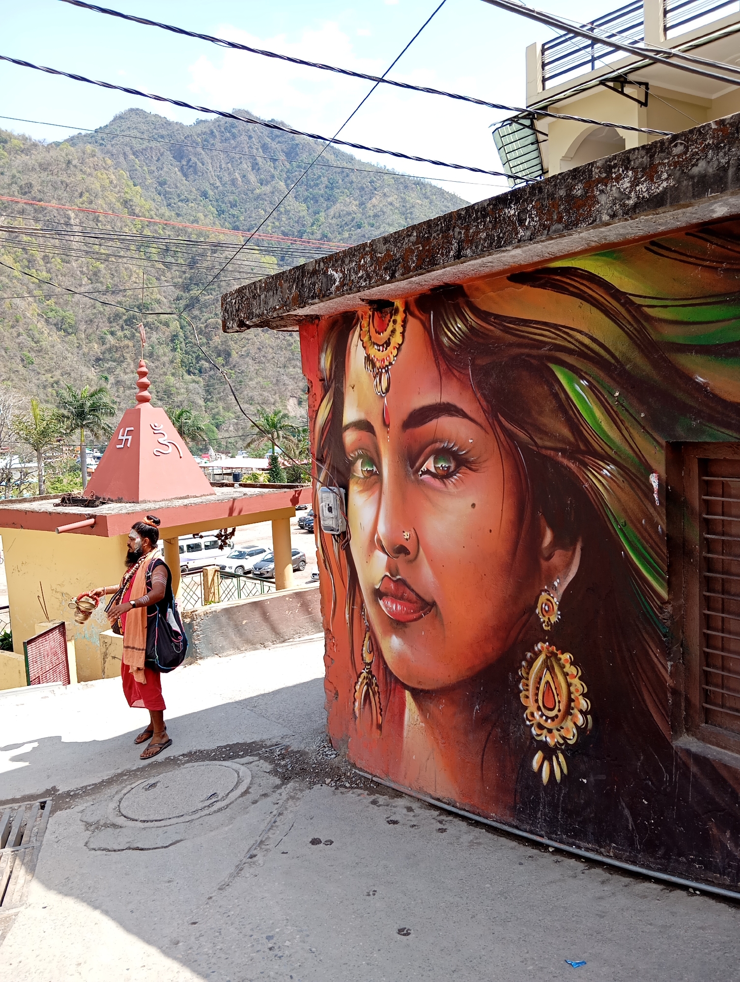 The image shows a street scene in Jonk, India. A man in traditional Hindu garb, with a beard and wearing saffron-colored clothing, is walking on a concrete street next to a small building. He is carrying a bag and what appears to be ritual items. The building has a large mural of a woman's face painted on its side. The mural depicts a woman with intricate jewelry and vibrant colors. Behind the building with the mural, there's a small shrine or temple with a pyramid-shaped roof. In the background, there is a mountain range and power lines overhead. Several cars are parked in a lot behind the shrine. The sky is clear and bright.