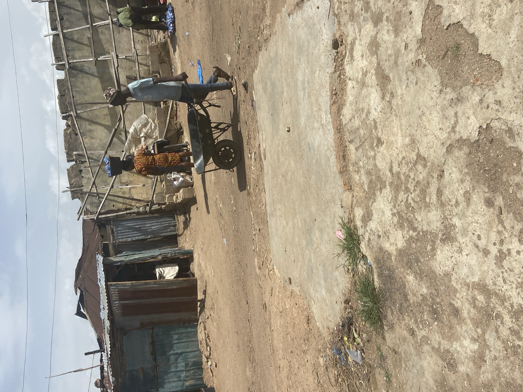 A man is pushing a wheelbarrow on a dirt road. He is wearing a blue t-shirt and jeans. He has on a patterned head covering and flip-flops. Behind him, a woman is walking with a bucket on her head. She is wearing a patterned dress. In the background, there are some buildings made of corrugated metal and wood. The sky is cloudy. This scene is in Bukuru, Nigeria.
