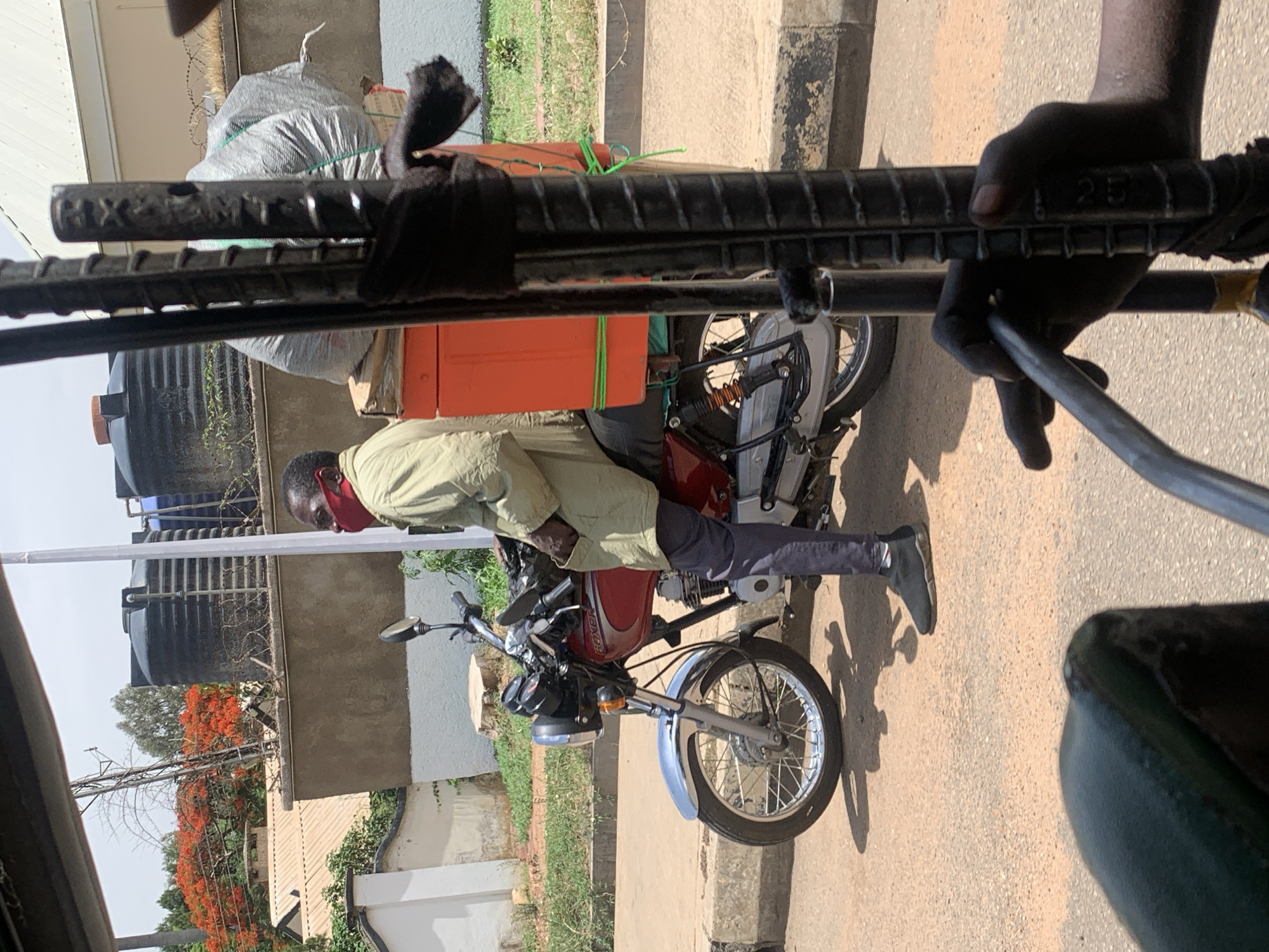 The scene captures a man in Jos, Nigeria, riding a motorcycle laden with goods. He wears a light green shirt, dark pants, and black shoes. A red face mask partially covers his face. The motorcycle is red and black, carrying a large orange box secured with green ropes, along with other wrapped packages. The rider seems to be seated on the motorcycle on the side of a paved street, with buildings and trees in the background. In the foreground are black metal bars, possibly part of a vehicle or structure, held by a dark-skinned hand.