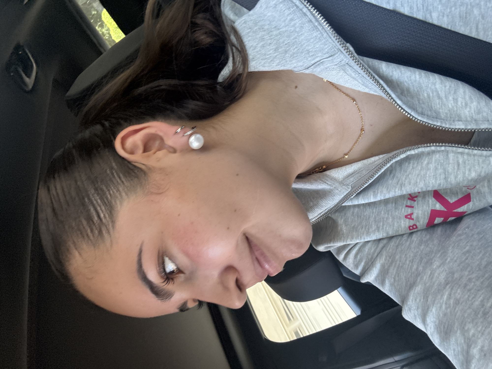 The image displays a close-up portrait of a young woman, rotated 90 degrees counter-clockwise, suggesting she is seated in a vehicle. She has dark hair pulled back and is looking downwards with a soft, subtle smile. Her left ear features a large white pearl earring in the lobe and two smaller silver hoop earrings higher up.
She is dressed in a grey hooded zip-up sweatshirt, and a delicate gold chain necklace with small beads adorns her neck. A visible mole is present on her upper left shoulder/neck area. A black seatbelt is stretched diagonally across her chest, confirming her location within a car.
The background is blurred, showing the dark interior of the vehicle, including a black seat or headrest. Through a window in the upper right, glimpses of blurred green foliage and possibly distant buildings are visible, indicating it is daytime. On the right side of her grey sweatshirt, a pink logo with the partial text "BAIK" and a large "K" is visible. The scene captures a casual moment in San Bernardino Tlaxcalancingo, Mexico.