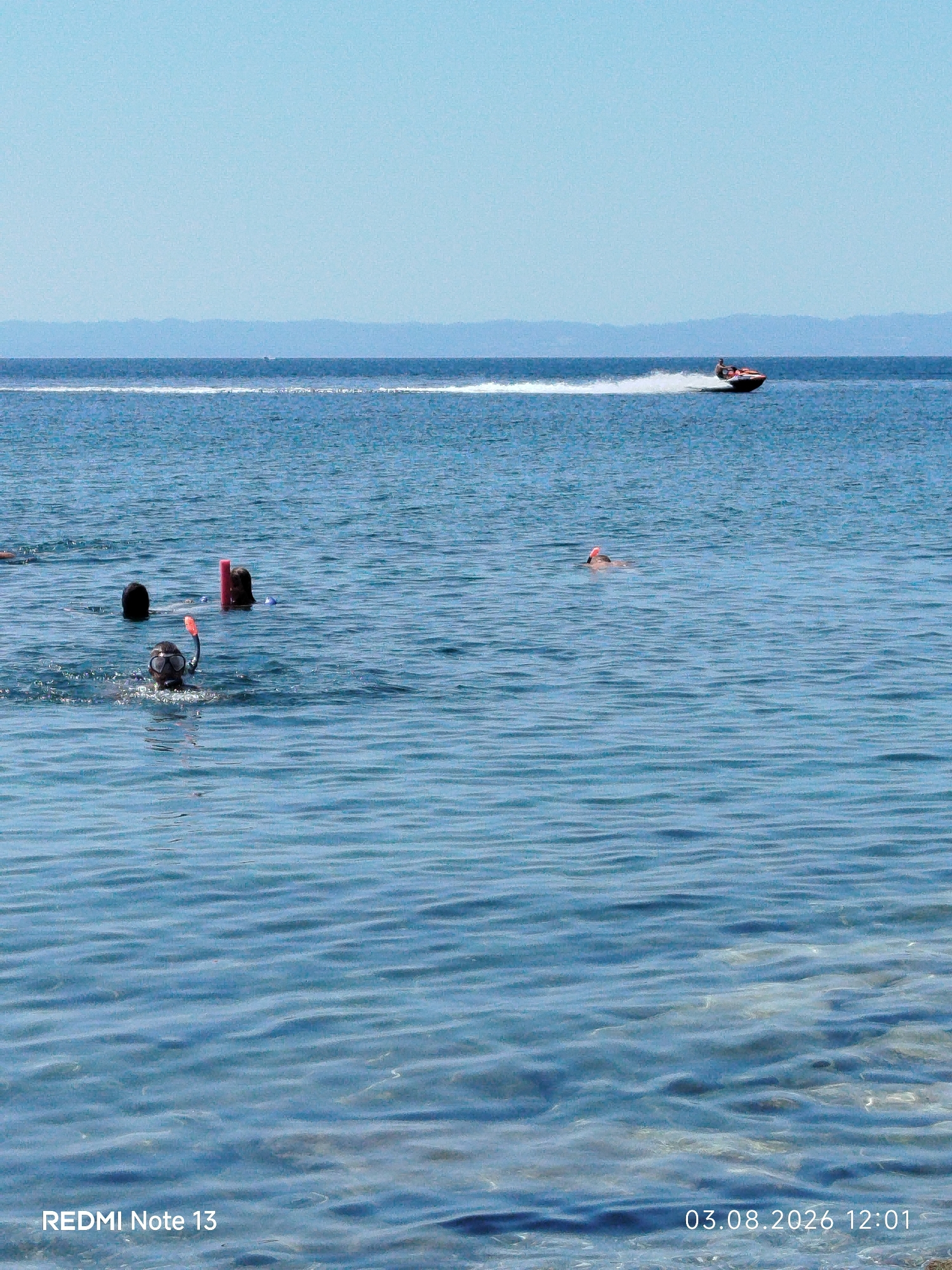 The image depicts a serene coastal scene in Sithonia Municipal Unit, Greece, under a clear blue sky. The foreground is dominated by the calm, blue-green waters of the Aegean Sea, with gentle ripples reflecting the sunlight. In the middle ground, several individuals are engaged in water activities. One person is snorkeling, wearing a mask and snorkel, with an orange buoy nearby. Two other individuals are partially submerged, with only their heads and shoulders visible. Further in the distance, a jet ski with two occupants is speeding across the water, leaving a white spray trail. The horizon line is marked by a distant landmass, likely the mainland of Greece or another island, with hazy, mountainous terrain. The overall atmosphere is one of leisure and enjoyment of a sunny day at the sea.