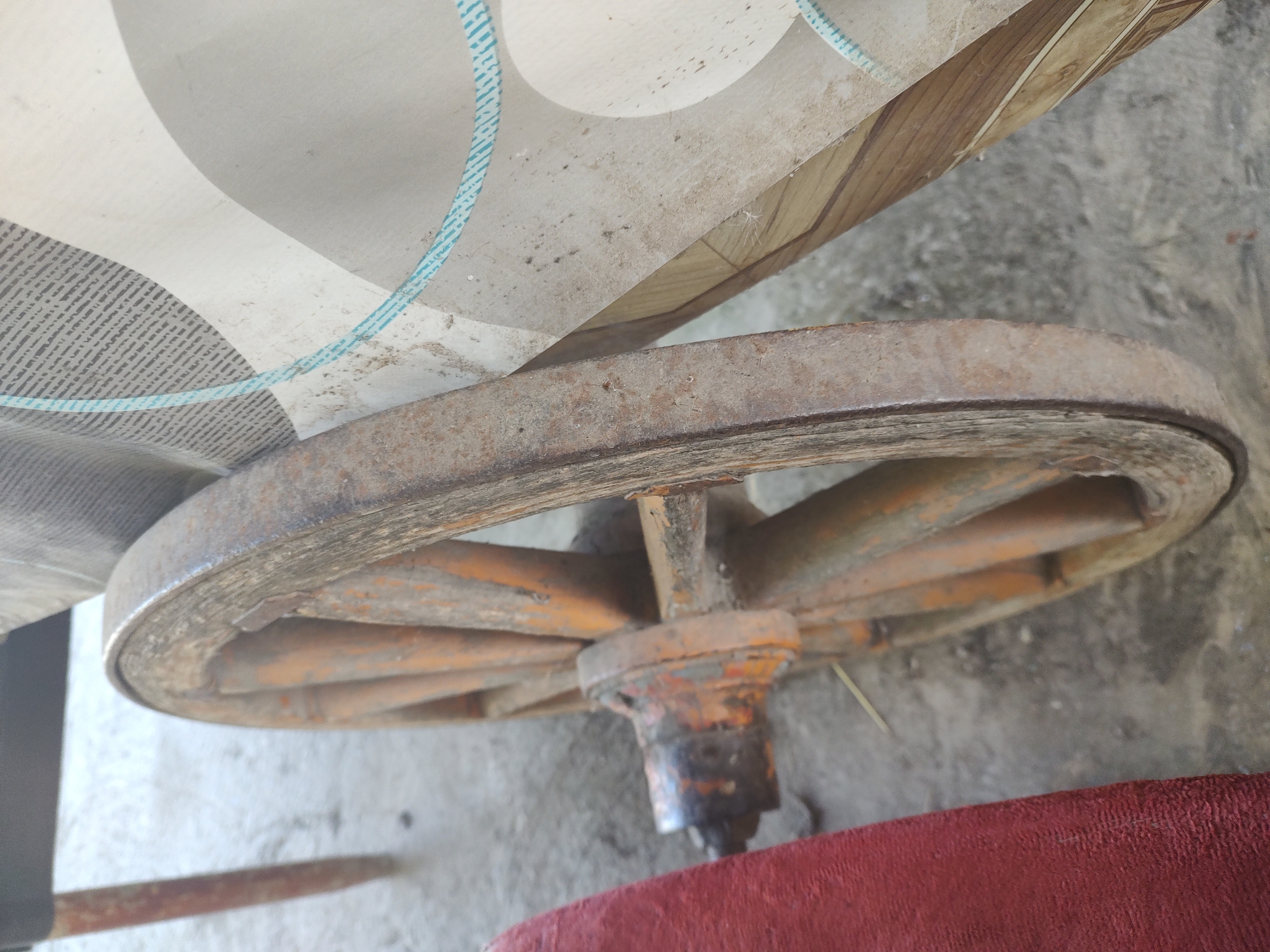 The image shows a close-up, angled view of an old, wooden wagon wheel. The wheel is weathered and appears to be made of several wooden spokes attached to a central hub, which is orange and rusted. The outer rim of the wheel is also worn and shows signs of rust and age. The wheel is resting on a concrete surface. To the left of the wheel, a section of a red upholstered object is visible. Behind the wheel, a patterned fabric or tarp is draped, featuring abstract shapes in shades of gray, white, and light blue stripes. The overall scene is outdoors, and the lighting suggests it is daytime. The context of Kochmar, Bulgaria is not visually discernible from the image itself.