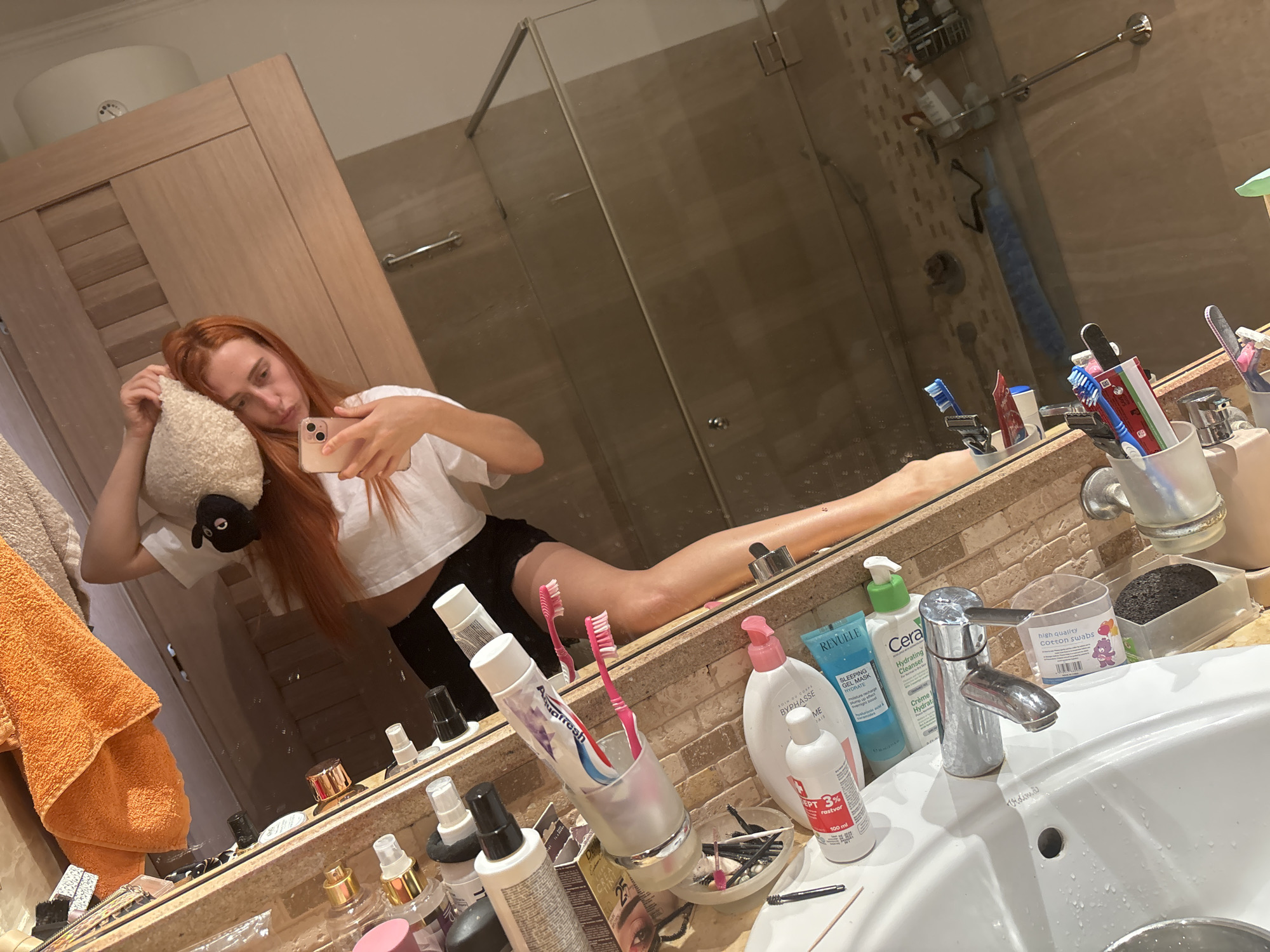 A young woman with long, auburn hair is taking a mirror selfie in a bathroom. She is wearing a white crop top and black shorts, and is holding a stuffed sheep toy. Her legs are extended in a split. The bathroom has a large mirror, a sink, and various toiletries on the counter, including toothbrushes, toothpaste, lotions, and cotton swabs. Visible text includes brand names like "Aquafresh," "BYPHASSE," "REVUELE," and "CeraVe," as well as product descriptions like "SLEEPING GEL MASK HYDRATE" and "HIGH QUALITY COTTON SWABS." There is also text in Cyrillic, indicating the location is Budva, Montenegro. The lighting suggests it is daytime.