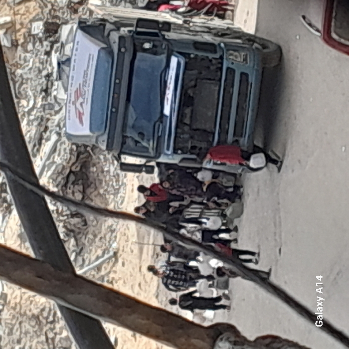 The scene in Khan Yunis, Palestinian Territory, depicts a humanitarian aid operation amidst a landscape of extensive rubble and demolition debris. A blue-grey semi-truck is centrally positioned on an unpaved dirt surface. The truck's cab features a white roof-mounted banner displaying "DRINKING WATER" and a red cross logo, consistent with Médecins Sans Frontières (MSF). The truck's front grille is absent, revealing internal engine components.
Approximately fifteen individuals, predominantly appearing to be children and young adults, are gathered around the truck. Several individuals are actively carrying white plastic containers or jugs, indicating water collection or distribution. Some people are seated on the open cargo area at the rear of the truck. One person in a red jacket is walking towards the right foreground, carrying a white container. Blurred, dark diagonal structural elements partially obscure the left side of the foreground.