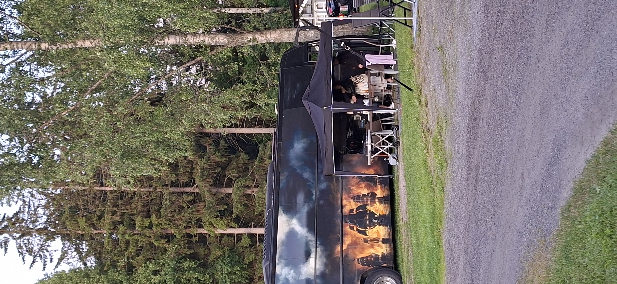 The image shows a black bus with a graphic of sailing ships on fire against a cloudy sky. In front of the bus is a black canopy tent set up over a table with chairs. Several people are standing and interacting under the tent, some holding cups or drinks. A portable grill and a gas cylinder are visible on the table. The scene is outdoors, with a gravel path and green grass in the foreground, and a dense forest of pine and birch trees in the background. There are also some white structures and chairs visible to the right of the canopy. The location appears to be a campsite or an outdoor event venue in Salpakangas, Finland.
