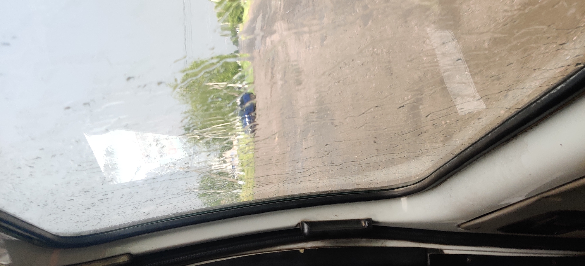 The image shows a view through a rain-streaked window of a vehicle, likely in Shelestove, Ukraine. Outside, a dirt road is visible, with a blue car parked on the side. Trees and some buildings can be seen in the background. The sky is overcast, indicating rainy weather. The interior of the vehicle, specifically the door and window frame, are visible on the left side of the image.