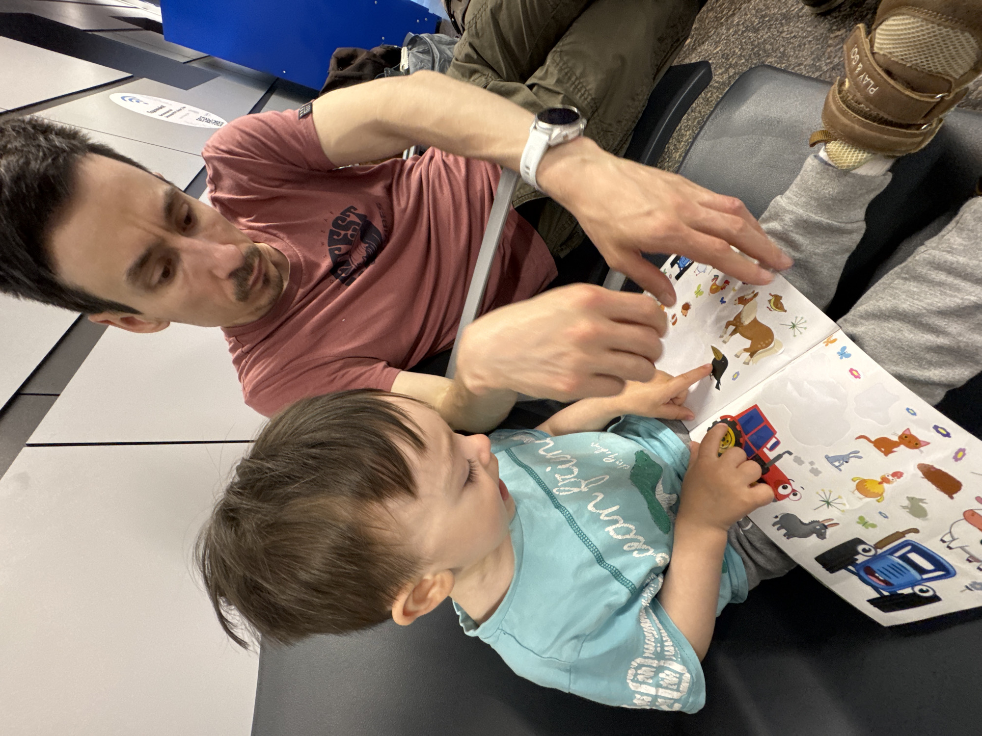 In an indoor setting, a man in a mauve-colored t-shirt with a graphic and a white smartwatch on his left wrist is seated and pointing at a sticker book. A young child wearing a light blue shirt with a green animal design is also seated and looking at the sticker book, holding a red toy. The sticker book contains images of various animals and vehicles. The man is likely the child's father. The scene appears to be in a waiting area, possibly an airport, as suggested by the chairs and wall panels. The city of Sochi, Russia is not discernible from the image.