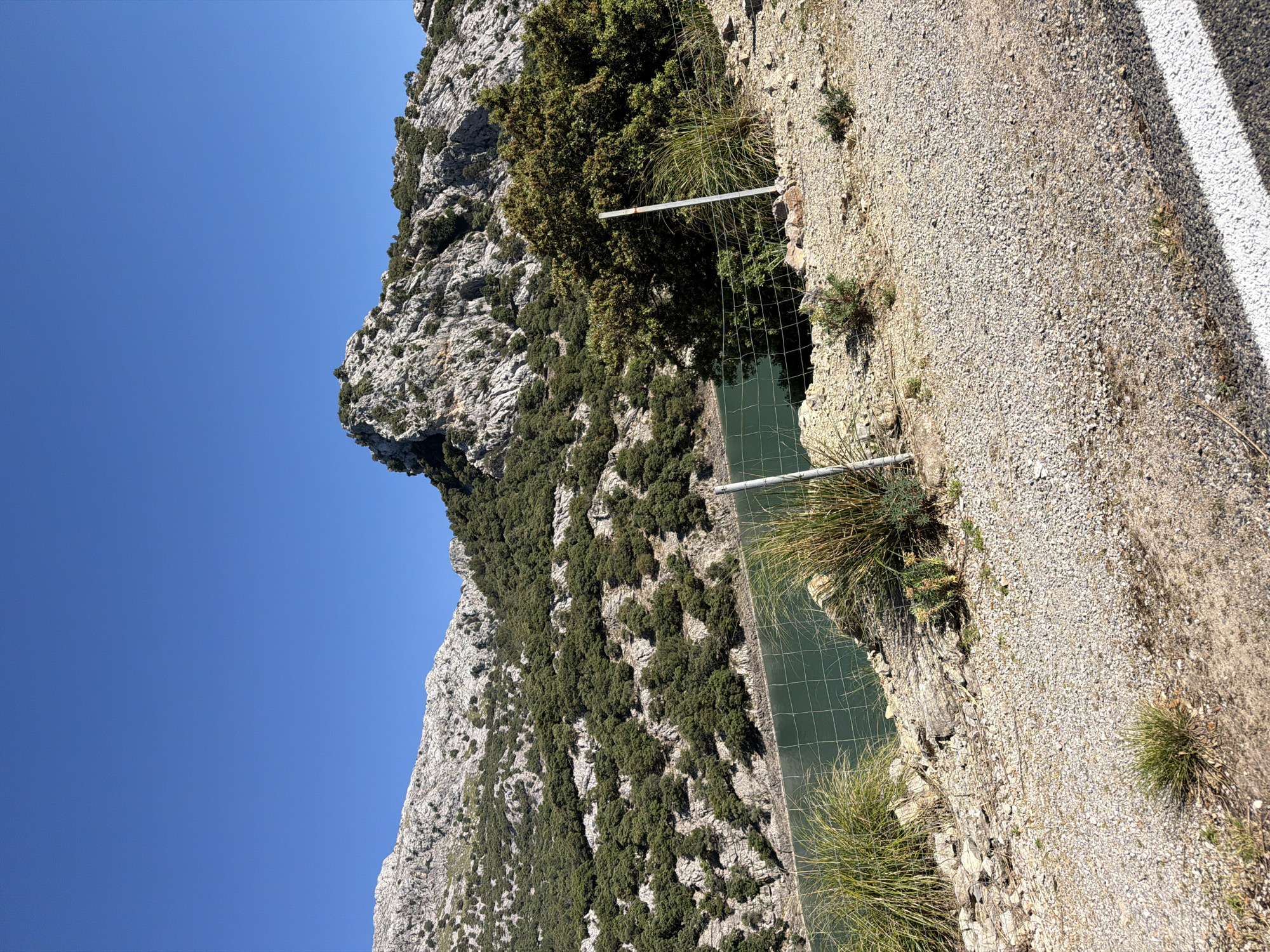 The image depicts a mountainous landscape in Escorca, Spain. A steep, rocky mountain covered with sparse trees and vegetation is visible on the left, set against a clear blue sky. To the right, a gravelly slope descends to a body of water, presumably a reservoir or lake, enclosed by a wire fence supported by metal posts. The edge of a paved road, marked with a white line, runs along the top right corner of the image.