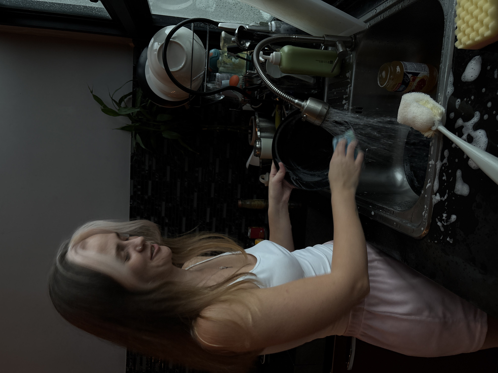 A woman with blonde hair and a white tank top and pink shorts is washing dishes in a kitchen. She is smiling and looking down at the pan she is scrubbing. There is a dish rack behind her with plates and glasses. The sink is full of soapy water and there are bubbles on the counter. The kitchen appears to be modern and clean.