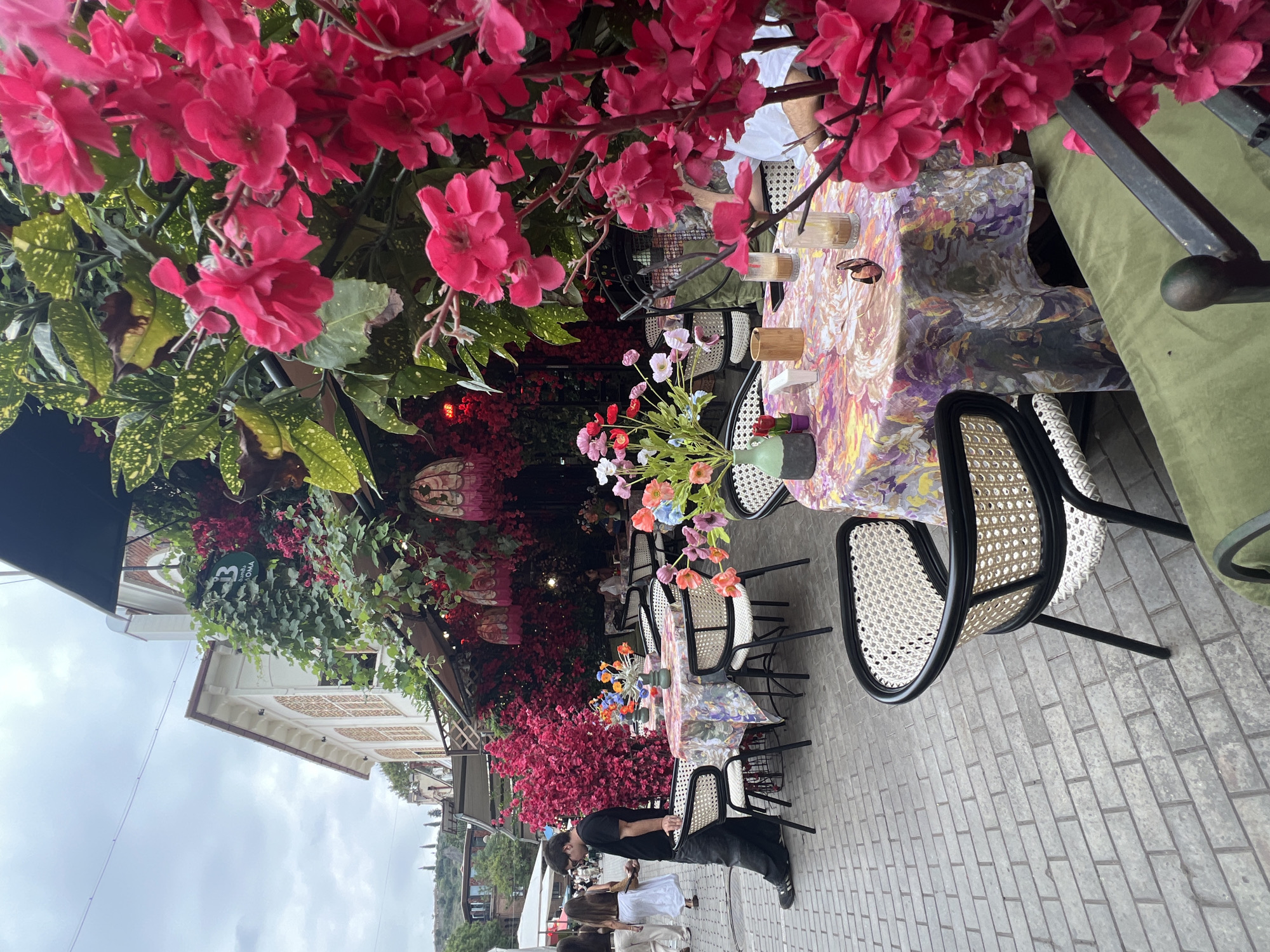This image depicts an outdoor seating area of a cafe or restaurant on a cobblestone street in Tbilisi, Georgia. The area is densely decorated with vibrant pink and red flowers, vines, and hanging lanterns, creating a lush, almost whimsical atmosphere. Several tables are set up with chairs, and one table in the foreground is covered with a colorful floral tablecloth and features a vase of flowers, glasses of drinks, and sunglasses. A man wearing a black t-shirt and jeans is standing near a table, looking at his phone. In the background, other patrons are visible, and a building with traditional Georgian architecture can be seen. The sky is overcast, suggesting a cloudy day.