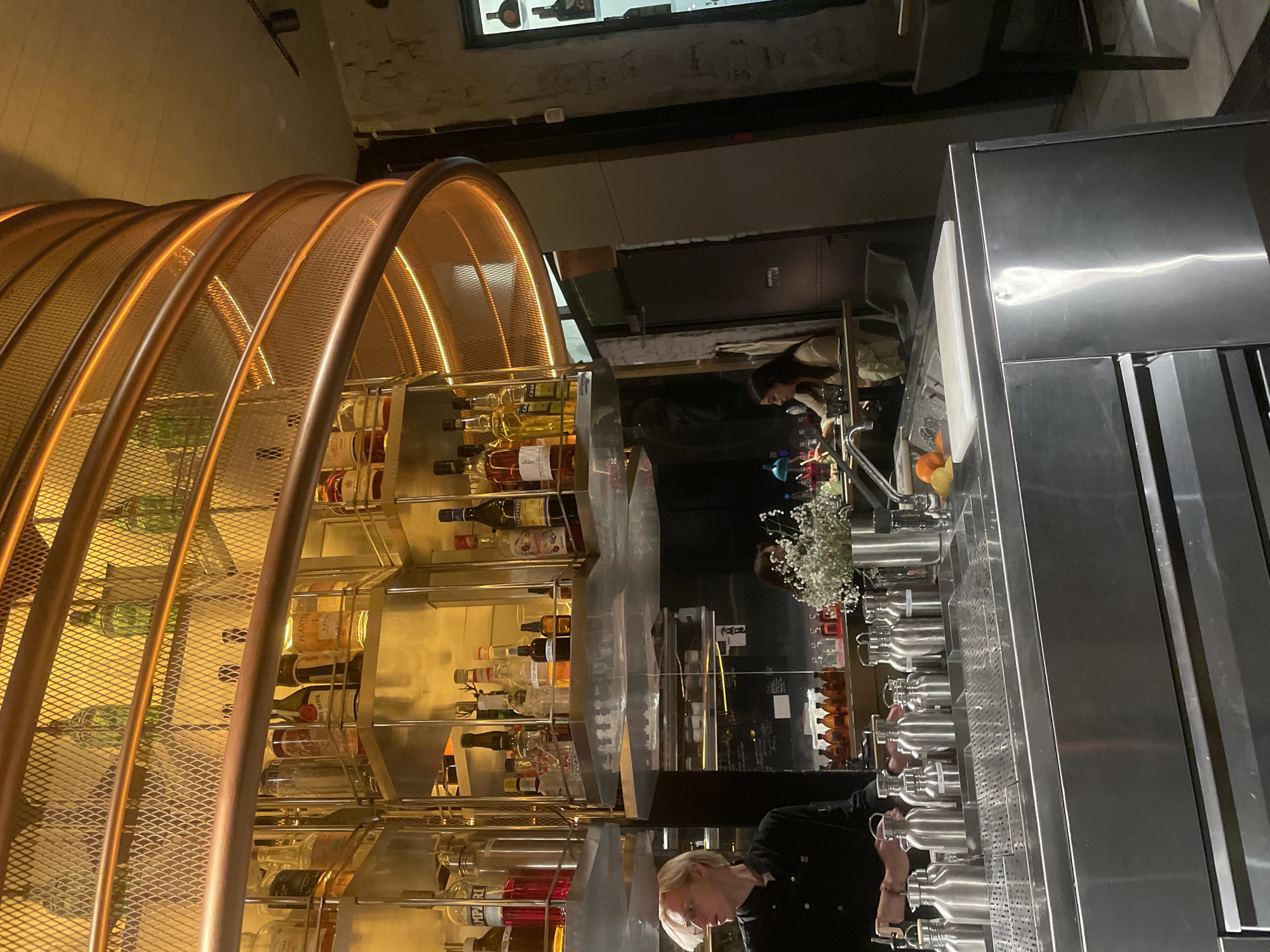 The image shows the interior of a bar, likely located in Tel Aviv, Israel, based on common bar styles in the city. The scene is dominated by a large, copper-colored metal structure that arches over the bar area, featuring a mesh design and internal lighting. Shelves behind the structure display various bottles of alcohol, creating a well-stocked bar appearance. A bar employee wearing a black shirt is visible, seemingly working on or near a stainless steel countertop lined with various fixtures and equipment, possibly related to dispensing or mixing drinks. There are stainless steel containers visible on the counter. A woman with long hair is visible by a counter that includes fruit. The environment appears to be relatively dimly lit, with the bar's lighting accentuating the copper structure and the bottles.