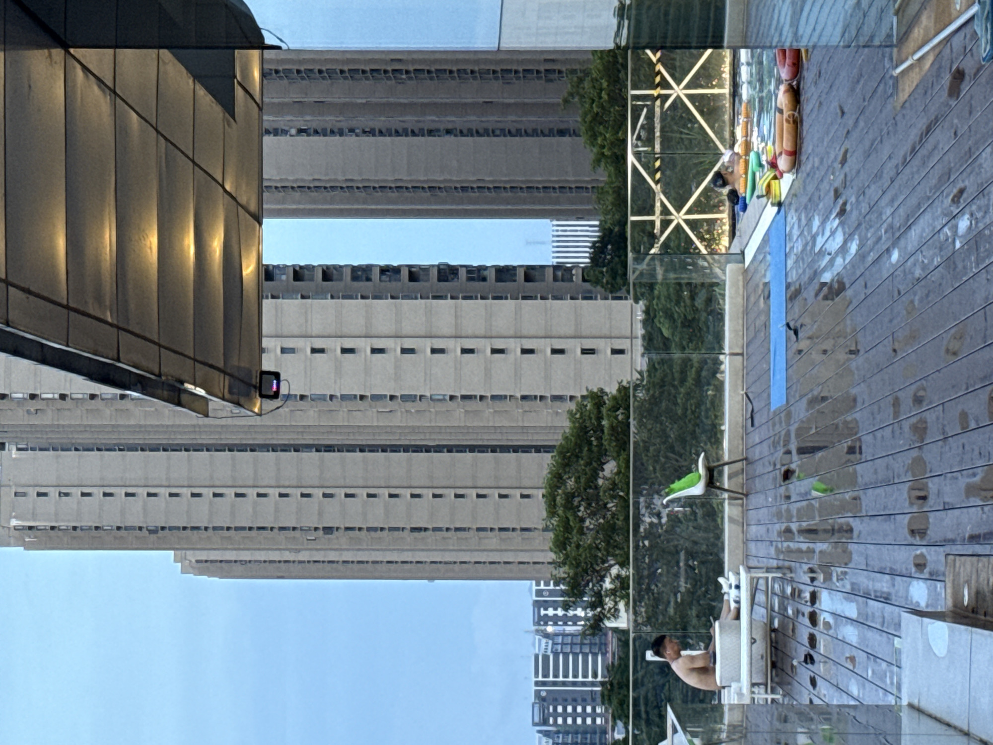 This media file captures a scene from a high-rise building with a view of other similar buildings and a swimming pool area. The image is oriented vertically, with the sky appearing at the bottom and the ground at the top.
In the foreground, a man is seated by a table on a wooden deck. He appears to be relaxed, with his upper body visible. The deck is wet, suggesting recent rain or cleaning. Nearby, there are chairs, one of which has a bright green cushion. A few inflatable rings, one orange and one yellow, are visible by the edge of the pool. The swimming pool itself has a blue lane marker.
In the background, towering concrete buildings dominate the view. They are characterized by numerous small, dark-framed windows arranged in rows. The architecture suggests a dense urban environment. Trees are visible between some of the buildings and behind the pool area, providing a touch of greenery.
The time of day appears to be late afternoon or early evening, as indicated by the warm, golden light reflecting off the angled glass facade of the building in the upper left portion of the image. The sky is a clear, light blue, suggesting fair weather. The overall impression is one of an urban oasis within a large city, offering a space for relaxation amidst the bustling metropolis. There is no visible text in the image.
