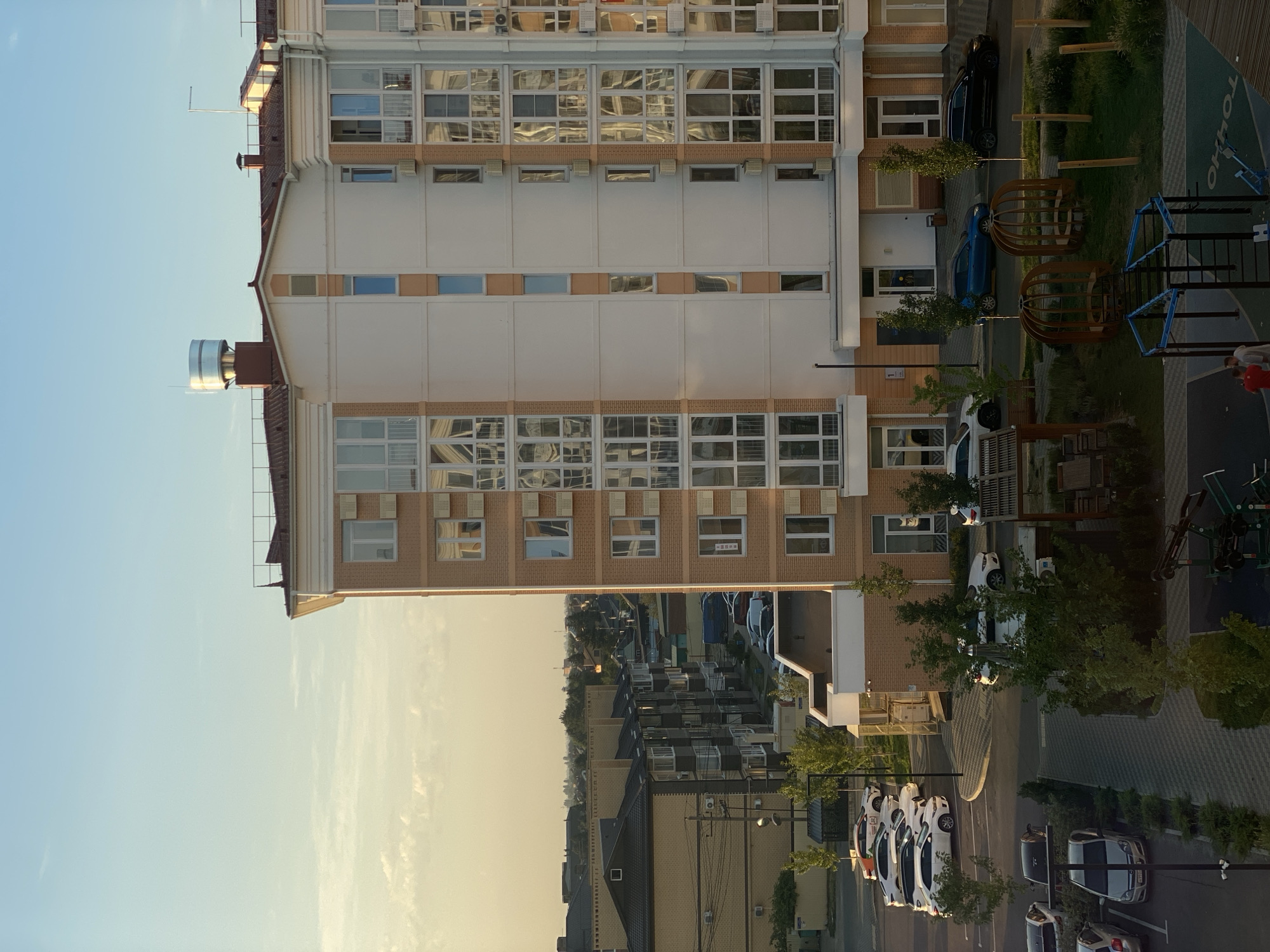 The image depicts an aerial view of a residential area in Agronom, Russia, likely during late afternoon or early evening given the warm, soft lighting. The scene is dominated by a modern, multi-story apartment building with a light-colored facade and brown brick accents, featuring numerous windows that reflect the sky and surrounding environment. In front of this building, a parking lot is filled with various cars, mostly white, suggesting a residential complex.
To the left of the apartment building, several other buildings are visible, including a lower, wider building with a dark roof and light brown brickwork, also appearing residential. The street in front of these buildings is lined with parked cars and trees. Further in the distance, a sprawling cityscape with smaller houses and buildings stretches to the horizon under a pale blue sky with scattered clouds.
In the foreground, to the right of the apartment building, there is a children's playground with blue and black equipment and wooden seating structures resembling birdcages. A few people, two men, are standing near the playground equipment, appearing to be in conversation. The ground is a mix of paving stones and grass. The overall impression is one of a modern, developing urban or suburban environment.