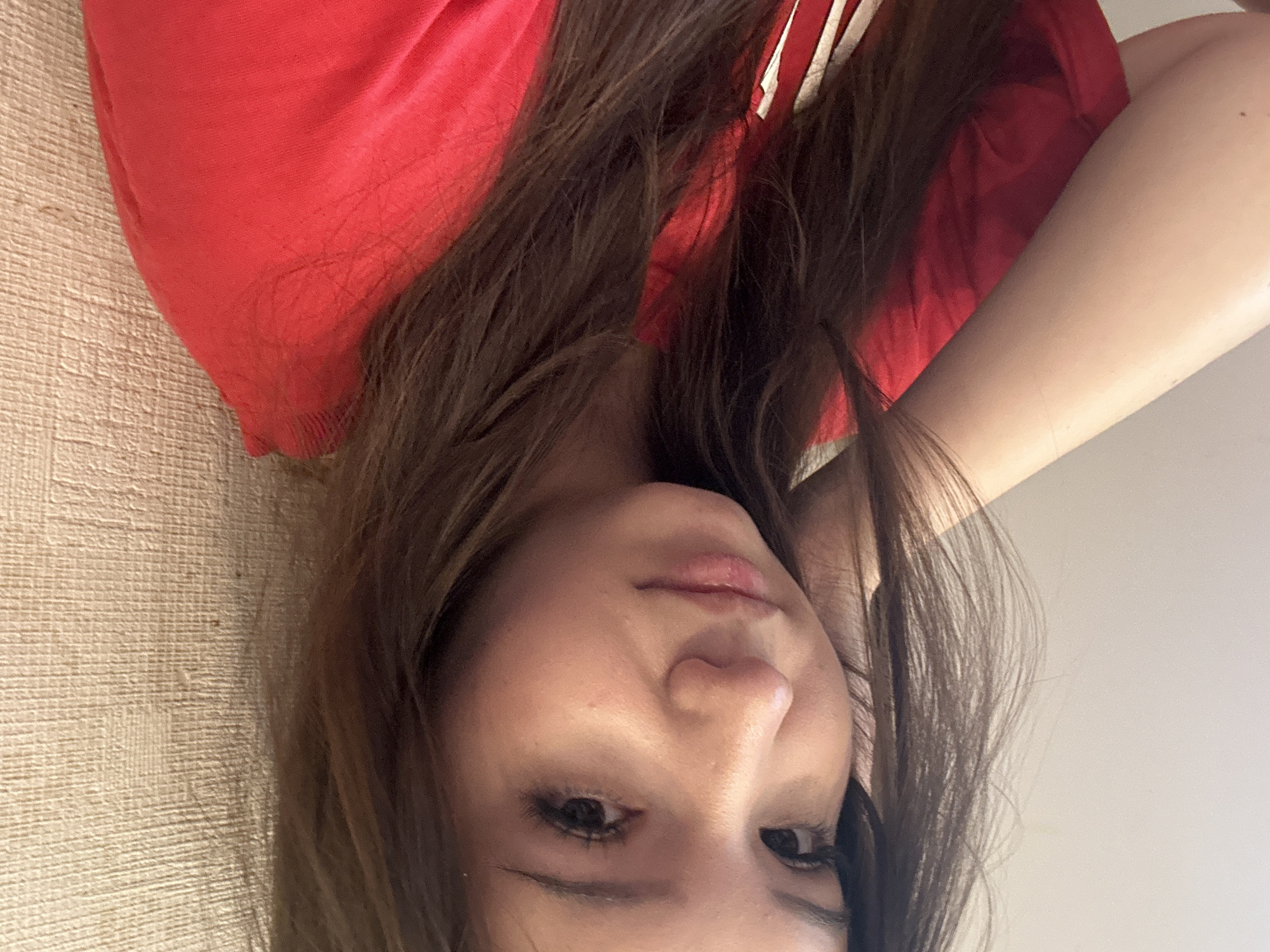 A young female, appearing to be in her late teens or early twenties, is depicted in a medium close-up, directly facing the camera with a neutral, slightly contemplative expression. She has long, wavy dark brown hair, parted slightly off-center, with some strands framing her face. Her dark eyes are open, and her full lips are closed. Her fair complexion shows minor visible pores around her nose and cheeks. She is wearing a short-sleeved, round-neck red t-shirt, possibly featuring a white stripe or logo on the lower left side (her right). Her left arm is bent, with her hand lightly touching the side of her face near her ear. The background consists of two distinct walls: a smooth, light beige or off-white wall on her left, and a textured, light tan wallpaper with a fine, woven pattern on her right. A small dark mark is visible on the textured wall. The scene is illuminated by soft, consistent indoor lighting. This scene is located in Naberezhnye Chelny, Russia.