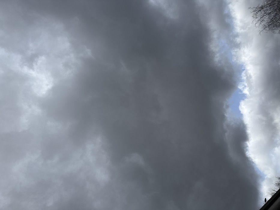 The scene depicts a sky heavily overcast with multiple layers of dark grey stratus and nimbostratus clouds, indicative of an impending or ongoing weather event. A narrow, irregular opening in the lower cloud layer, positioned towards the bottom-center, reveals a small segment of lighter grey clouds and a sliver of blue sky. In the bottom-left quadrant, a dark, angular structure consistent with a roofline or eave is partially visible. To the bottom-right, the bare, leafless branches of a deciduous tree extend into the frame against the lighter clouds. No individuals, explicit actions, or interactions are present. The environmental conditions suggest a day with low visibility and potential precipitation in Oberhausbergen, France.