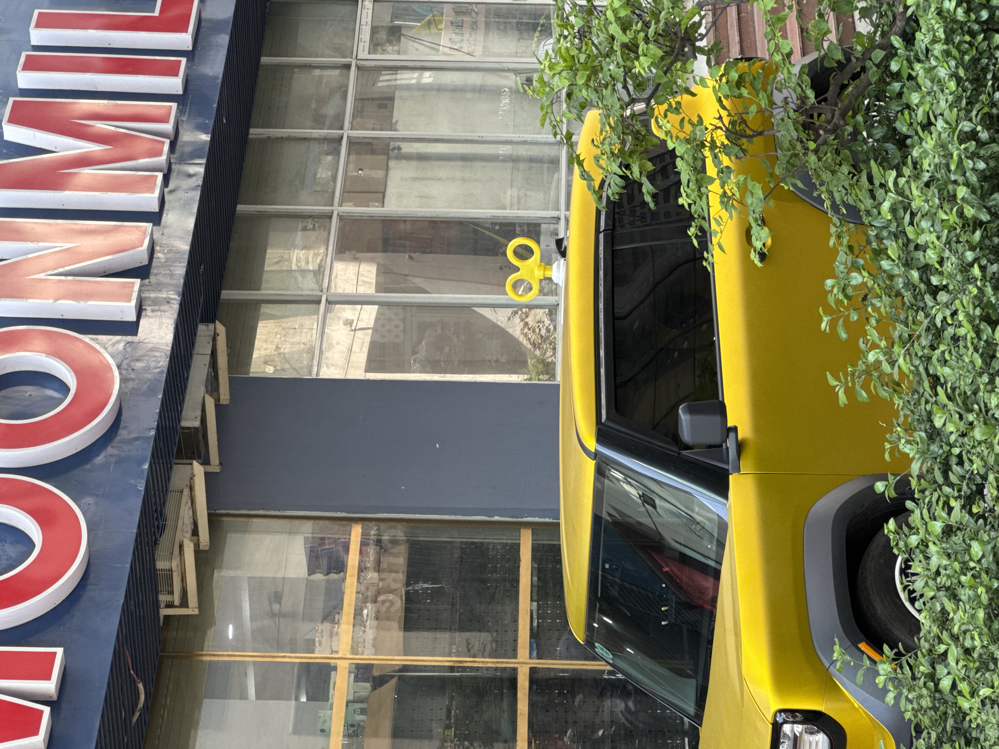 In Bac Nha Trang Ward, Vietnam, a bright yellow car is parked in front of a building. The building has large windows and a blue facade with large red letters spelling out what appears to be a store name. On the roof of the car, a yellow wind-up key toy is attached. Lush green foliage partially obscures the lower portion of the car and the lower parts of the building. The environment suggests an urban setting, possibly a street or a commercial area. The overall scene is static, with no people visible or any apparent action taking place.