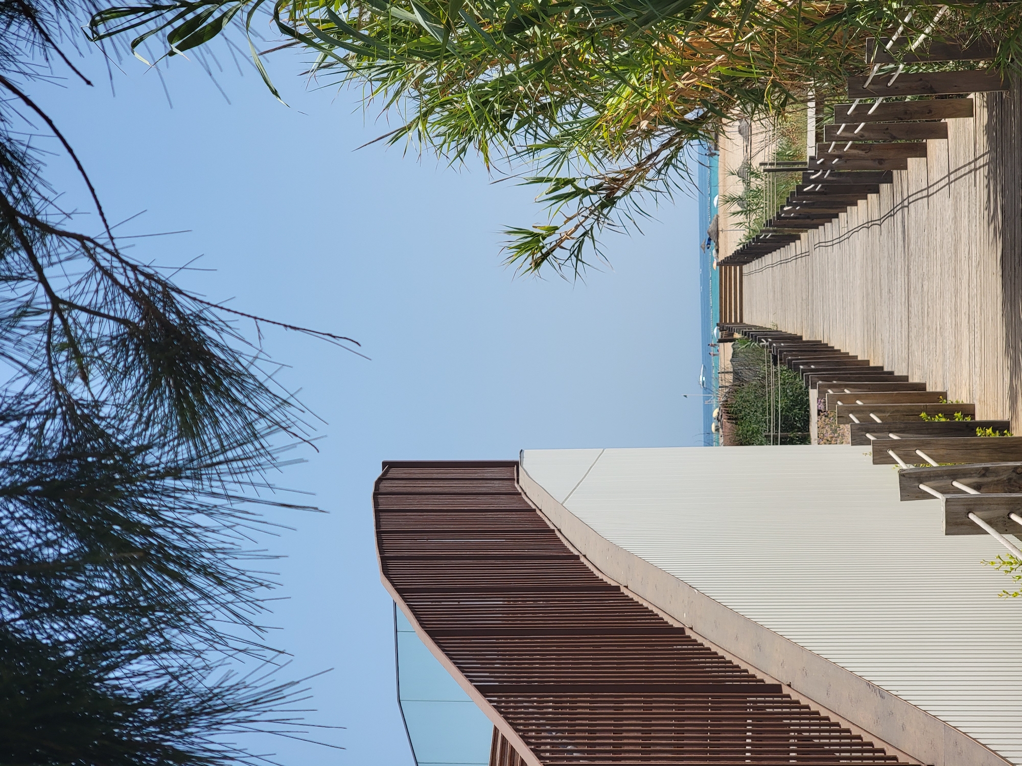 A wooden boardwalk extends towards a sandy beach and the ocean under a clear blue sky. On the left, a modern white building with vertical paneling is visible, featuring a curved wooden railing and glass barriers leading up a ramp. To the right, lush green vegetation grows along the boardwalk. In the distance, the beach is dotted with a few people and beach umbrellas, and a boat is visible on the calm water. The scene depicts a pathway to the beach in Quarteira, Portugal.