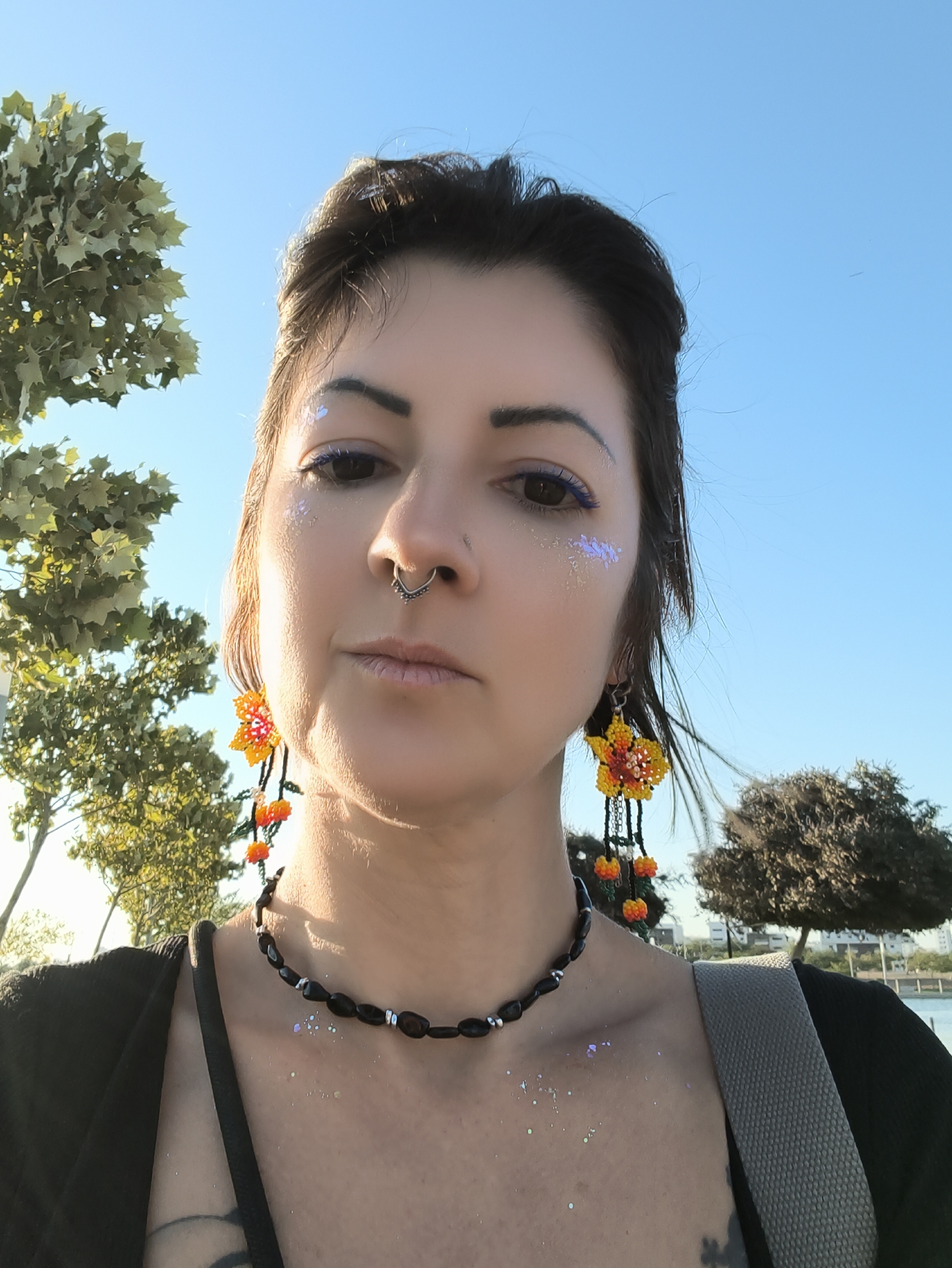 A young adult female is posing for a selfie outdoors. She has dark hair, a septum piercing, and is wearing large, colorful floral earrings and a black beaded necklace. There is glitter on her face and collarbone. She is wearing a black top with a grey strap across her shoulder, possibly part of a bag. Behind her, there are trees, a clear blue sky, and a body of water with buildings in the distance, suggesting a location like the Be'er Sheva River Park. The lighting indicates it is daytime, possibly late afternoon, with the sun casting a warm glow.