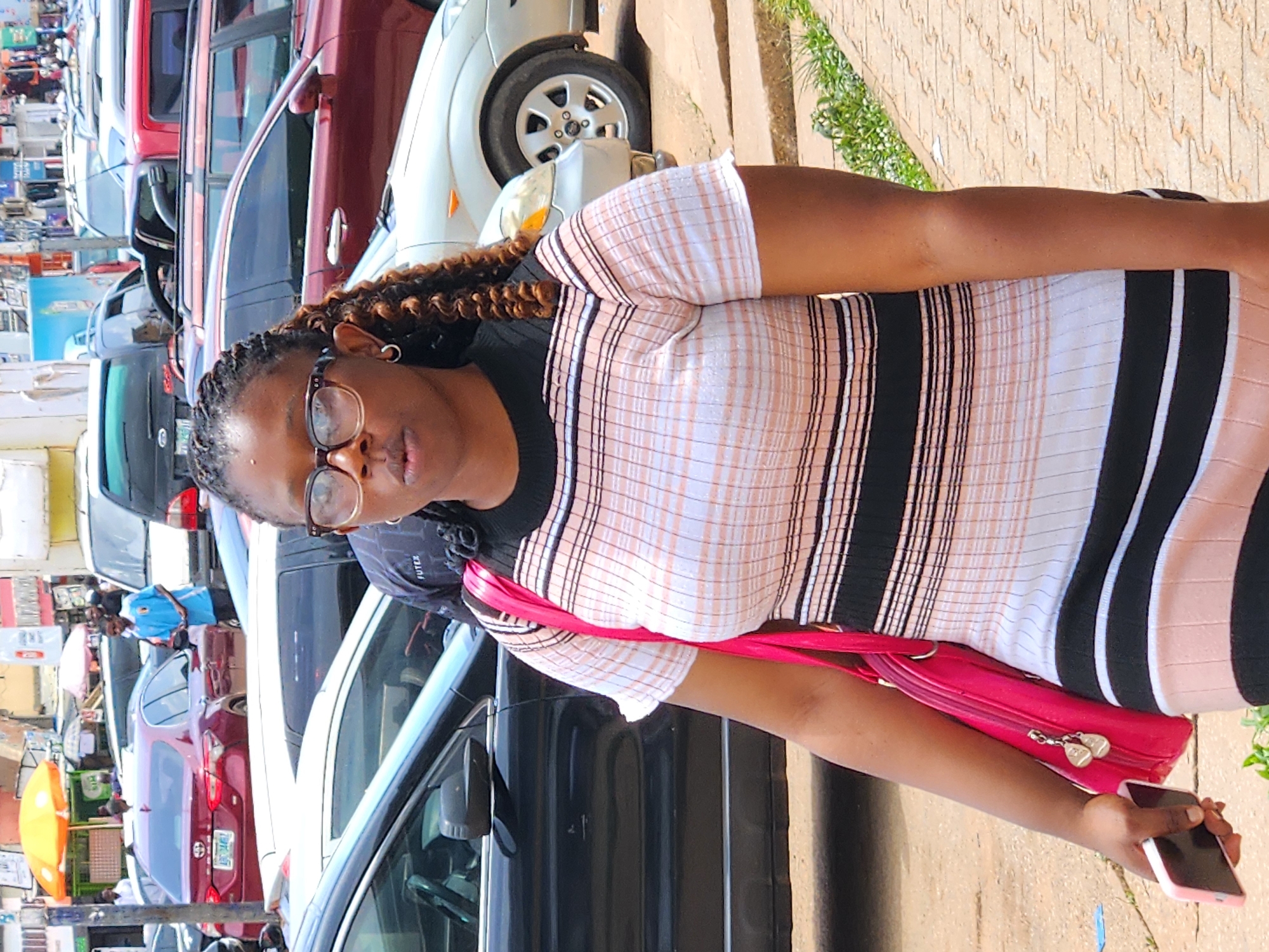 An outdoor, eye-level, medium shot shows a woman of African descent standing in the foreground on a sidewalk in what appears to be a busy urban environment. The woman is wearing a black, white, and light pink horizontally striped, short-sleeved, fitted dress and glasses. Her dark hair is braided with some strands falling over her shoulders. She is holding a pink smartphone in her left hand and a bright pink handbag over her left shoulder. Her right hand is not visible. In the background, multiple cars are parked or driving on a street, including a red sedan, a dark-colored SUV, and a silver hatchback. The environment is sunny, with a paved sidewalk and a dirt/gravel area. Buildings and streetlights are visible in the far background. The overall scene suggests a daytime street scene in a city.