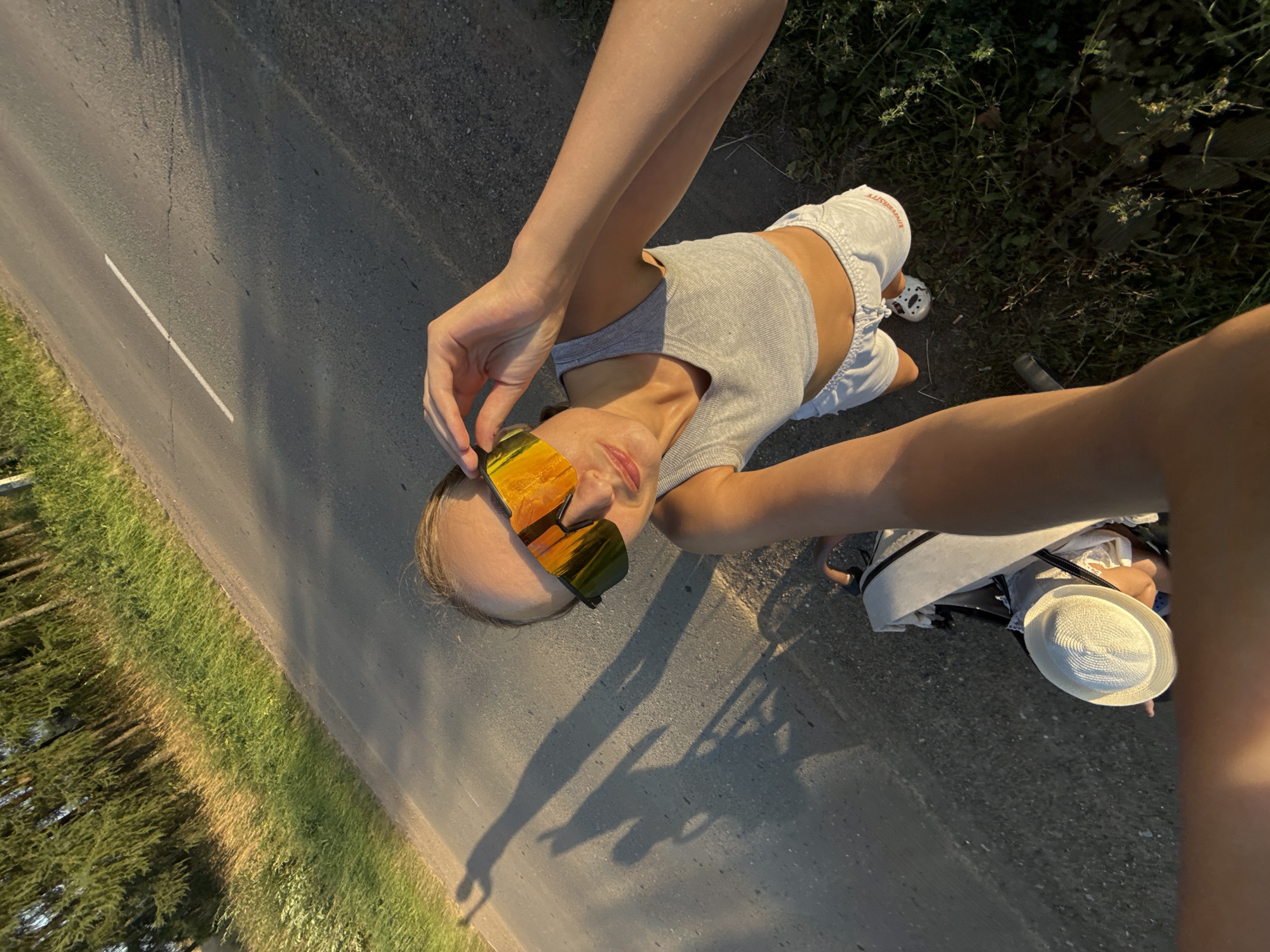 A young woman with blonde hair, wearing mirrored sunglasses, a gray crop top, and white shorts, is posing for a selfie outdoors on a sunny day. She is standing next to a stroller containing a baby wearing a white hat. The woman is adjusting her sunglasses with her right hand. They are on a paved road with a white line in the center, bordered by a grassy verge and trees. Shadows of people and possibly a bicycle are visible on the road. The scene appears to be located in the vicinity of a road in the village of Podshivalovo, Russia.