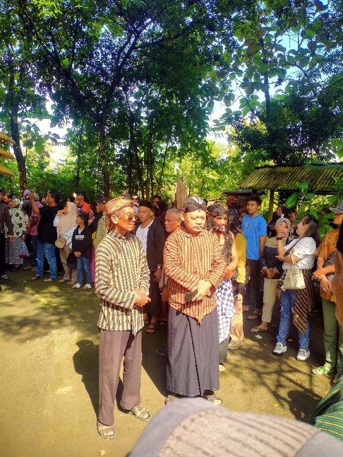 The scene is a gathering of people in Masaran, Indonesia, located at latitude -7.471, longitude 110.957. The scene is outdoors, with trees and foliage surrounding a paved area. The gathering includes many people, some wearing traditional clothing, including men with sarongs and women with scarves. Two men in the foreground stand facing the camera with hands clasped in front of them. They are wearing traditional Indonesian clothing, and are the focus of the scene. The other people in the group are standing, some with hands clasped in front of them, and some with their hands at their sides. They are observing the man in the foreground.