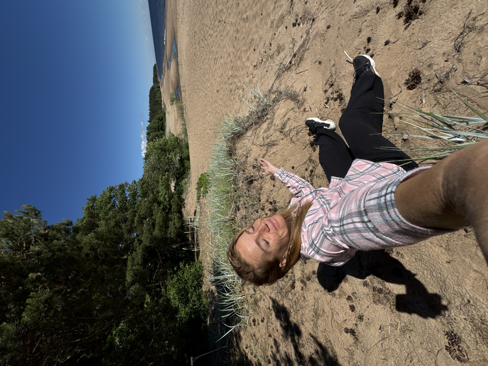 A woman with blonde hair and freckles is lying on a sandy beach, holding a phone in her right hand and taking a selfie. She is wearing a pink and white plaid shirt and black pants. Her eyes are closed and she is smiling. The beach is lined with trees and the sky is blue. The location is Zelenaya Roshcha, Russia.