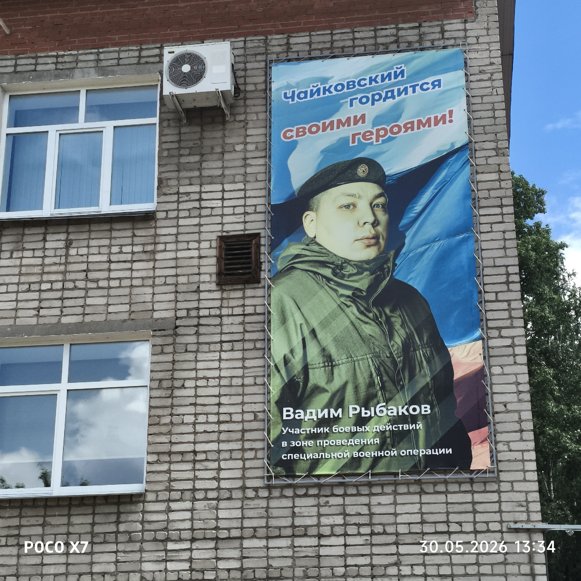 The image depicts a banner on a brick building, presumably in Chaikovsky, Russia, as indicated by the text on the banner. The banner displays a portrait of a man in military attire, identified as Vadim Rybakov, a participant in combat operations in the zone of the "special military operation." Above the portrait, the text reads "Chaikovsky is proud of its heroes!" The building has two white-framed windows and an air conditioning unit on the wall. The image also includes a timestamp of "30.05.2026 13:34" and the text "POCO X7", which appears to be from the camera that took the picture. Trees are visible to the right of the building.