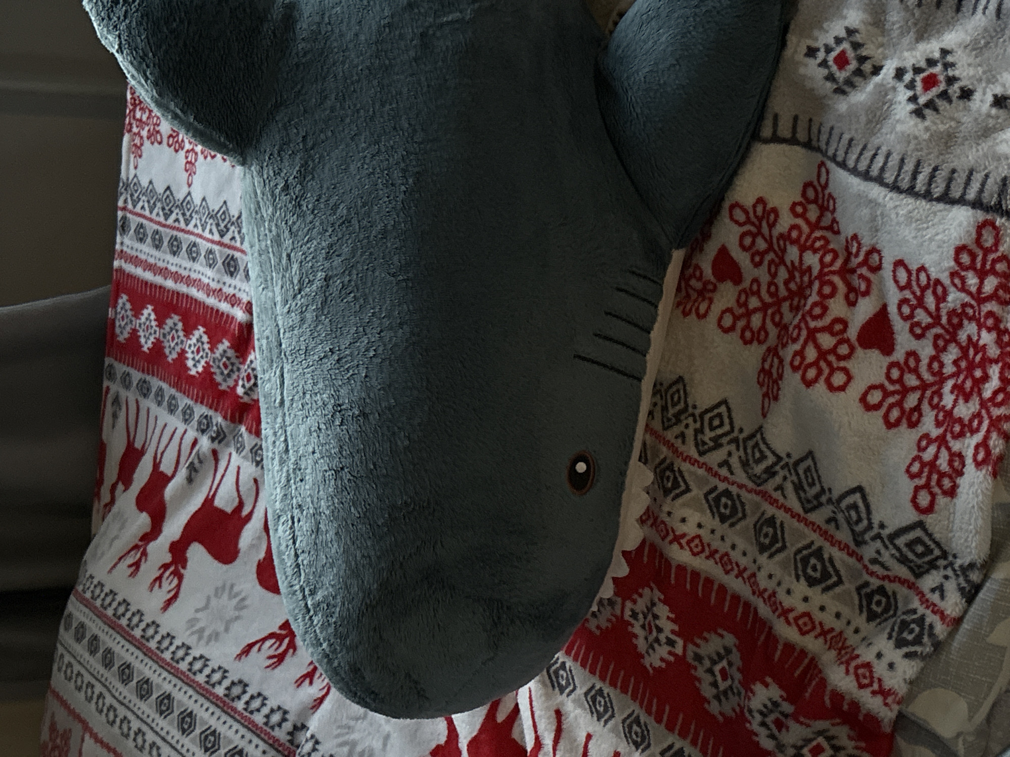 A plush blue shark toy rests on a patterned blanket with a red and white holiday-themed design featuring reindeer, snowflakes, and geometric shapes. The blanket is draped over what appears to be a bed or soft surface. The lighting is indoor and diffused. The location is not discernible from the image.